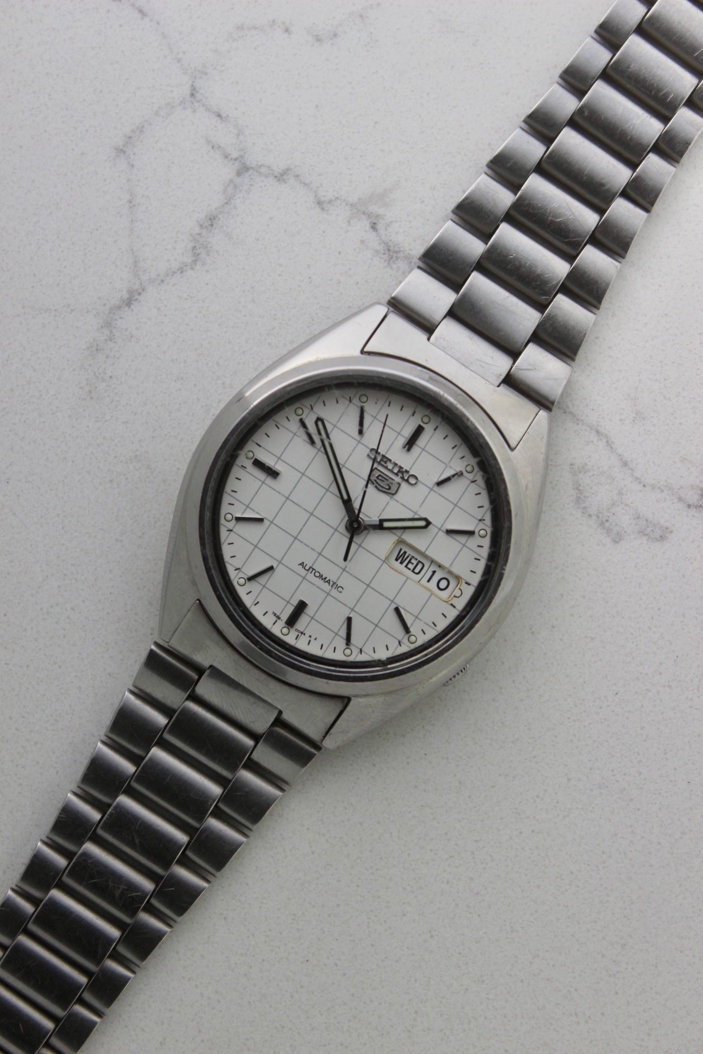 A vintage Seiko 5 automatic wristwatch (Reference Number: 7S26-3040) with a stainless steel case and bracelet. The watch features a 36mm white dial with a grid-painted design and a day-date function at the 3 o’clock position. The stainless steel bezel is fixed, complementing the original bracelet, which fits up to a 7-inch wrist. This Japan-exclusive mid-1980s model is in good condition, showing signs of wear on the bracelet, buckle, and crystal.