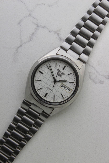A vintage Seiko 5 automatic wristwatch (Reference Number: 7S26-3040) with a stainless steel case and bracelet. The watch features a 36mm white dial with a grid-painted design and a day-date function at the 3 o’clock position. The stainless steel bezel is fixed, complementing the original bracelet, which fits up to a 7-inch wrist. This Japan-exclusive mid-1980s model is in good condition, showing signs of wear on the bracelet, buckle, and crystal.