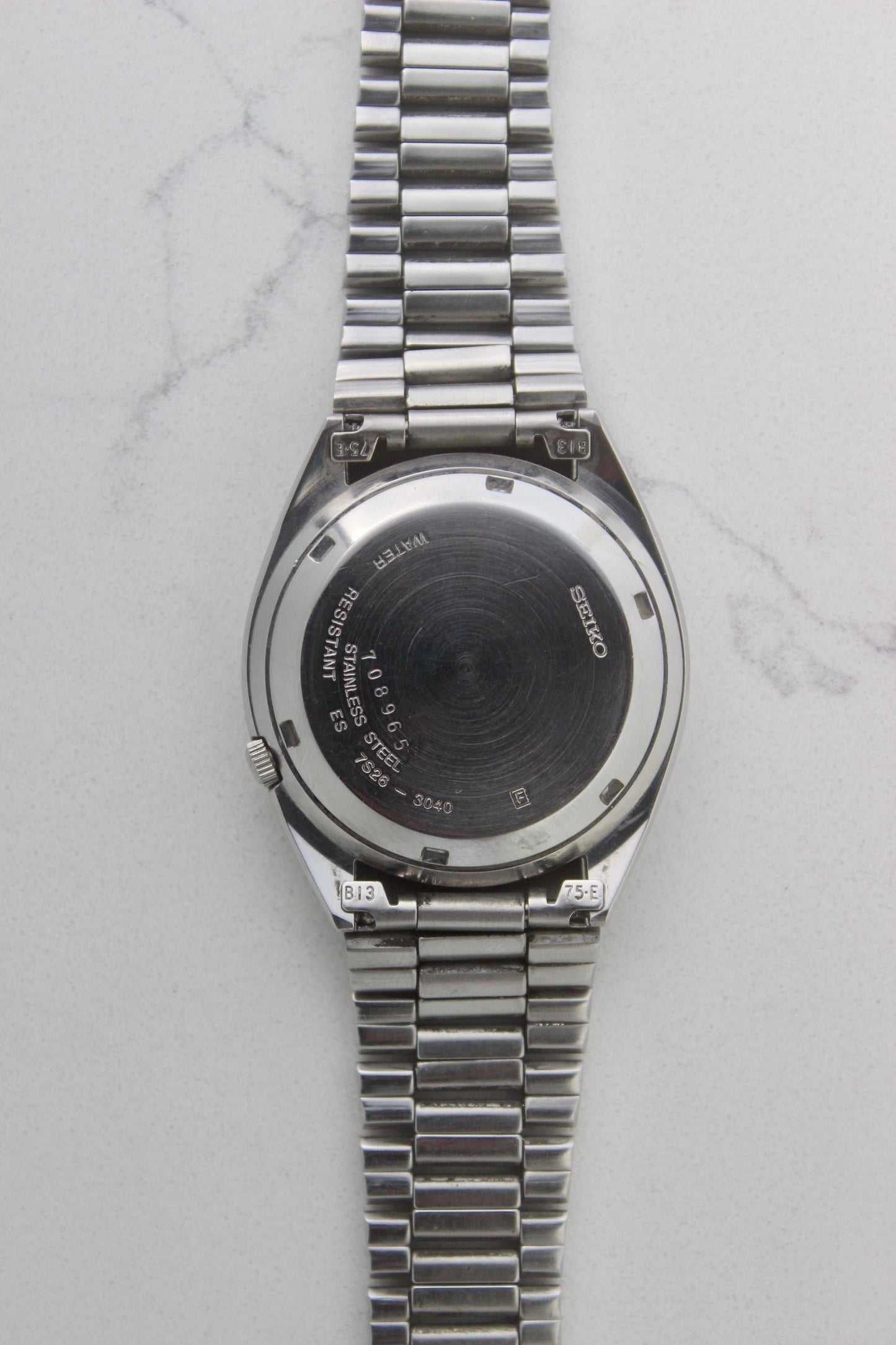 The case back of a vintage Seiko 5 automatic wristwatch (Reference Number: 7S26-3040) made of stainless steel. The screw-down case back is engraved with Seiko branding, water resistance details, and serial numbers, showing visible signs of wear including surface scratches. The original stainless steel bracelet is attached, featuring end links marked "B13" and "75-E". This Japan-exclusive mid-1980s model is in good condition with expected wear for its age.