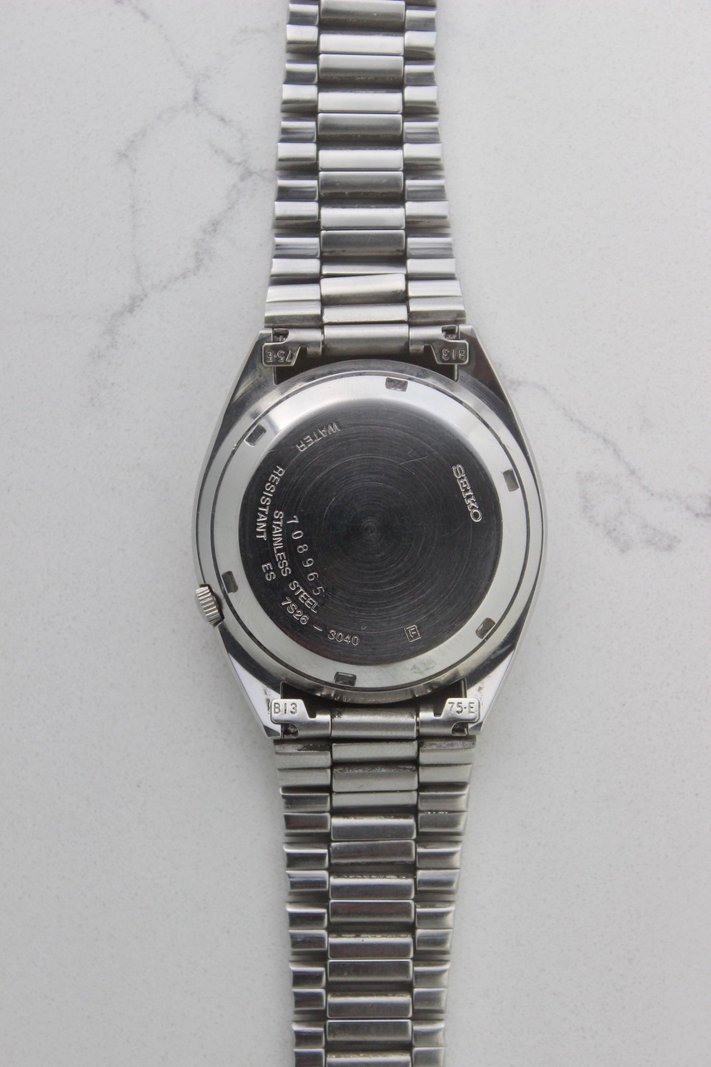 The case back of a vintage Seiko 5 automatic wristwatch (Reference Number: 7S26-3040) made of stainless steel. The screw-down case back is engraved with Seiko branding, water resistance details, and serial numbers, showing visible signs of wear including surface scratches. The original stainless steel bracelet is attached, featuring end links marked "B13" and "75-E". This Japan-exclusive mid-1980s model is in good condition with expected wear for its age.