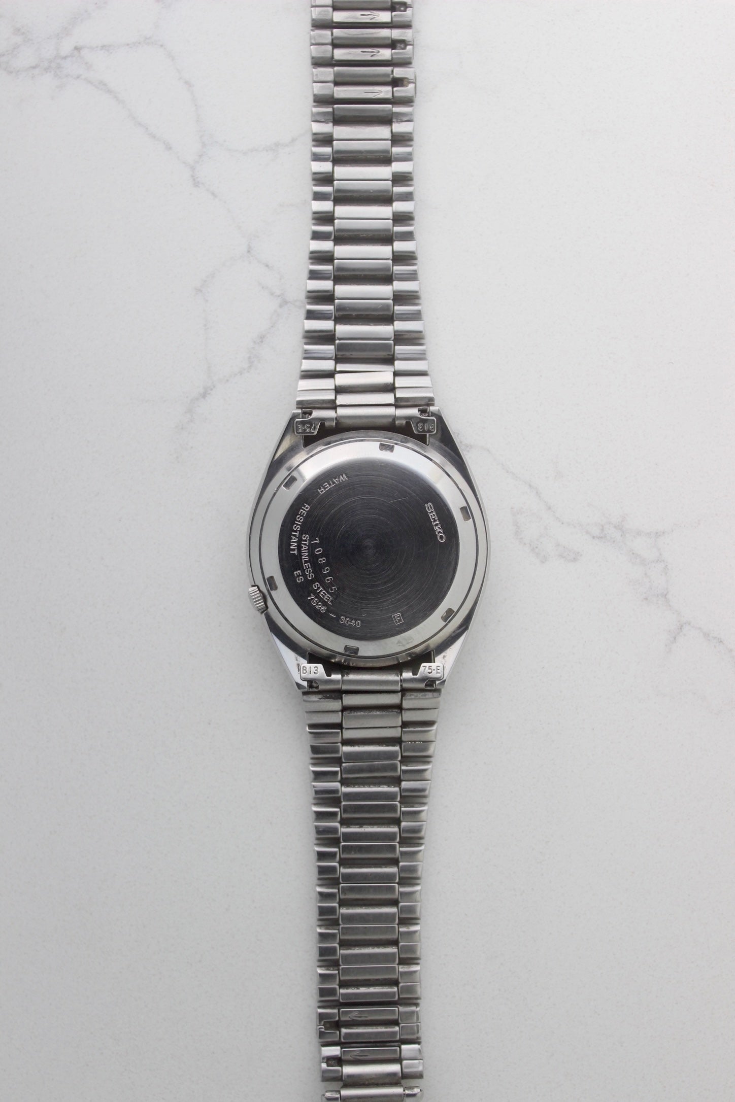 The case back of a vintage Seiko 5 automatic wristwatch (Reference Number: 7S26-3040) made of stainless steel. The screw-down case back is engraved with Seiko branding, water resistance details, and serial numbers, showing visible signs of wear including surface scratches. The original stainless steel bracelet is attached, featuring end links marked "B13" and "75-E". This Japan-exclusive mid-1980s model is in good condition with expected wear for its age.