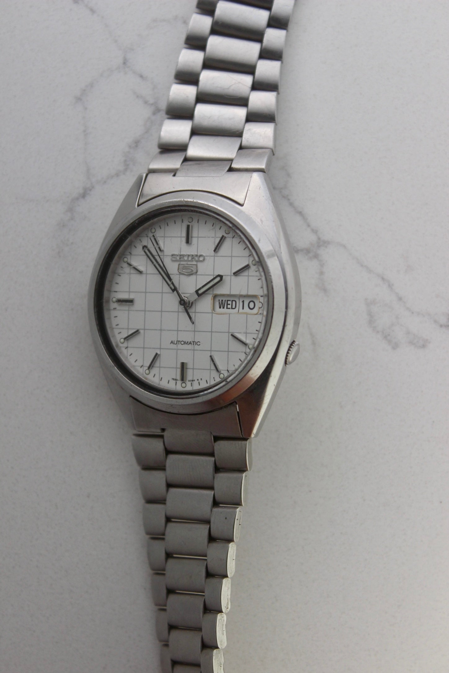  A vintage Seiko 5 automatic wristwatch (Reference Number: 7S26-3040) with a stainless steel case and bracelet. The watch features a 36mm white dial with a grid-painted design and a day-date function at the 3 o’clock position. The stainless steel bezel is fixed, complementing the original bracelet, which fits up to a 7-inch wrist. This Japan-exclusive mid-1980s model is in good condition, showing signs of wear on the bracelet, buckle, and crystal.