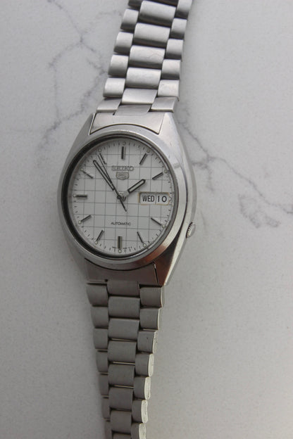  A vintage Seiko 5 automatic wristwatch (Reference Number: 7S26-3040) with a stainless steel case and bracelet. The watch features a 36mm white dial with a grid-painted design and a day-date function at the 3 o’clock position. The stainless steel bezel is fixed, complementing the original bracelet, which fits up to a 7-inch wrist. This Japan-exclusive mid-1980s model is in good condition, showing signs of wear on the bracelet, buckle, and crystal.