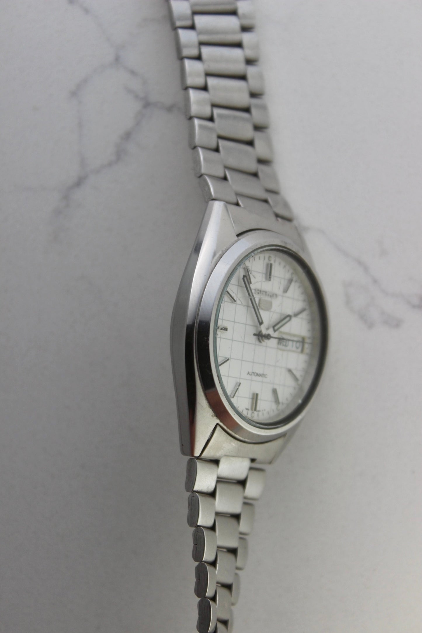 A vintage Seiko 5 automatic wristwatch (Reference Number: 7S26-3040) with a stainless steel case and bracelet. The watch features a 36mm white dial with a grid-painted design and a day-date function at the 3 o’clock position. The stainless steel bezel is fixed, complementing the original bracelet, which fits up to a 7-inch wrist. This Japan-exclusive mid-1980s model is in good condition, showing signs of wear on the bracelet, buckle, and crystal.