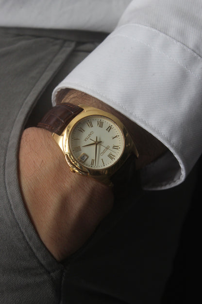 A detailed front view of the Seiko Caster
8F32-0140 watch featuring a gold-tone case, Roman numeral markers, and a date window at 3 o'clock. The cream-colored dial is complemented by classic black hands, encapsulating an elegant vintage aesthetic.
Paired with a genuine brown leather strap.