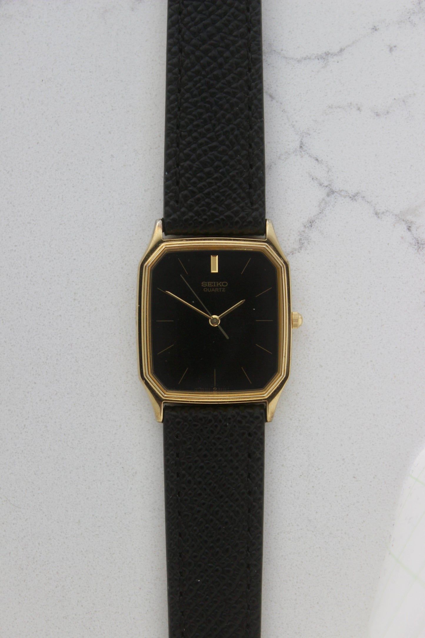 A close-up of the Seiko Chariot 69021-5180
featuring a sleek rectangular gold-plated case, a black dial with minimalist gold indices, and delicate gold-tone hands. The modern yet timeless design is paired with a handmade black leather strap stitched with beige accents for a refined look.