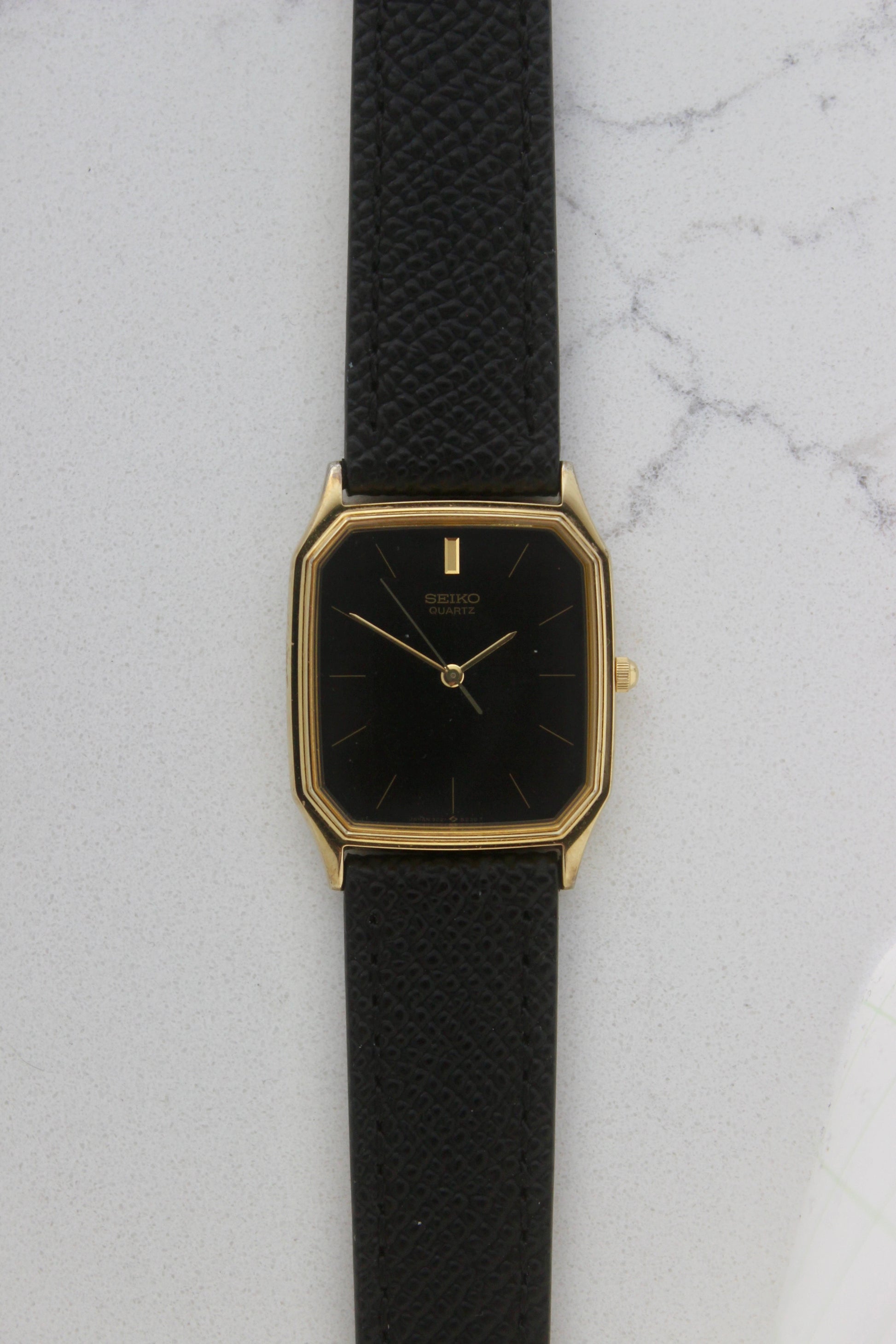 A close-up of the Seiko Chariot 69021-5180
featuring a sleek rectangular gold-plated case, a black dial with minimalist gold indices, and delicate gold-tone hands. The modern yet timeless design is paired with a handmade black leather strap stitched with beige accents for a refined look.
