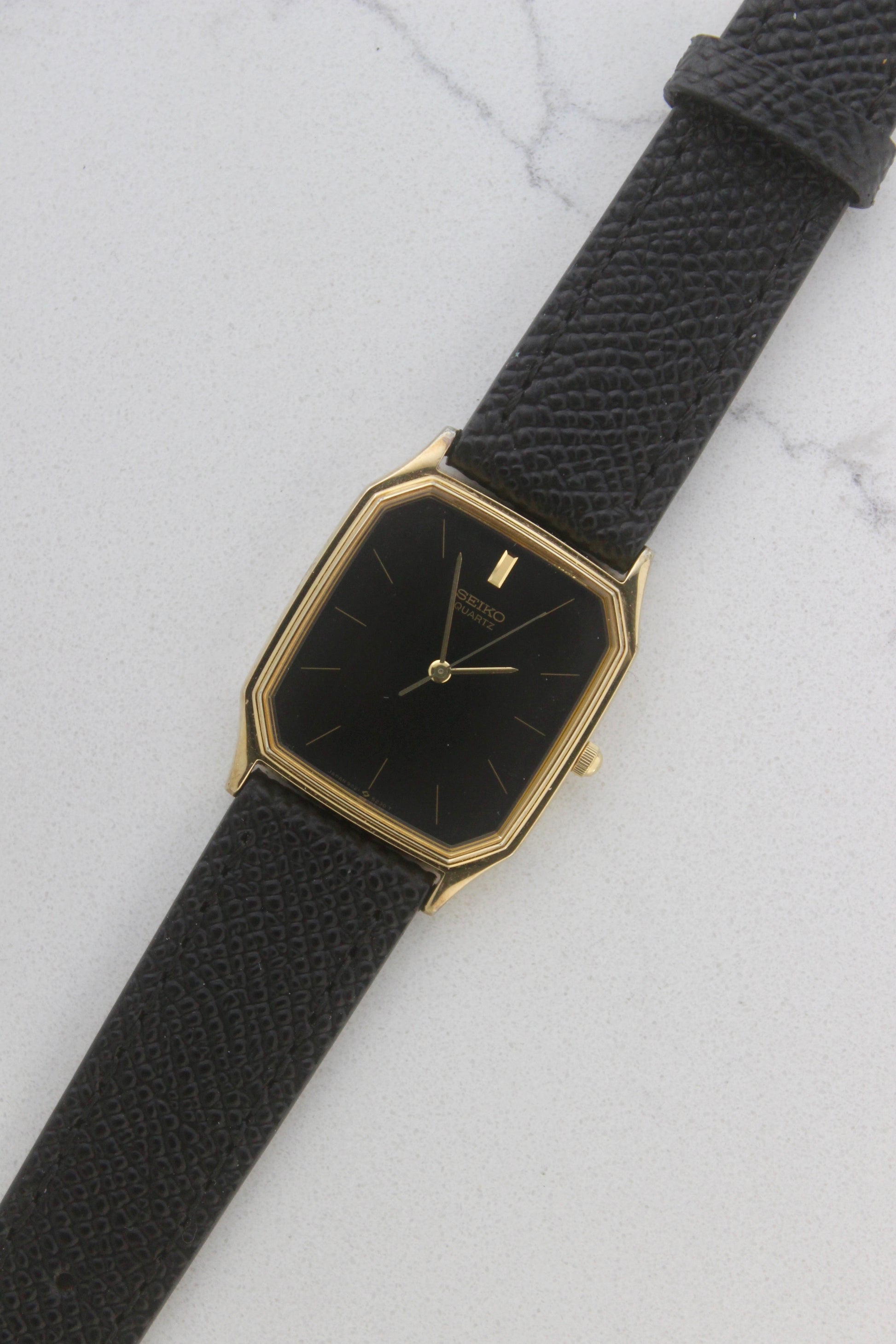 A close-up of the Seiko Chariot 9021-5180
featuring a sleek rectangular gold-plated case, a black dial with minimalist gold indices, and delicate gold-tone hands. The modern yet timeless design is paired with a handmade black leather strap stitched with beige accents for a refined look.