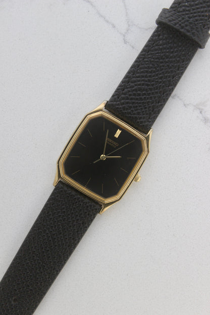 A close-up of the Seiko Chariot 9021-5180
featuring a sleek rectangular gold-plated case, a black dial with minimalist gold indices, and delicate gold-tone hands. The modern yet timeless design is paired with a handmade black leather strap stitched with beige accents for a refined look.