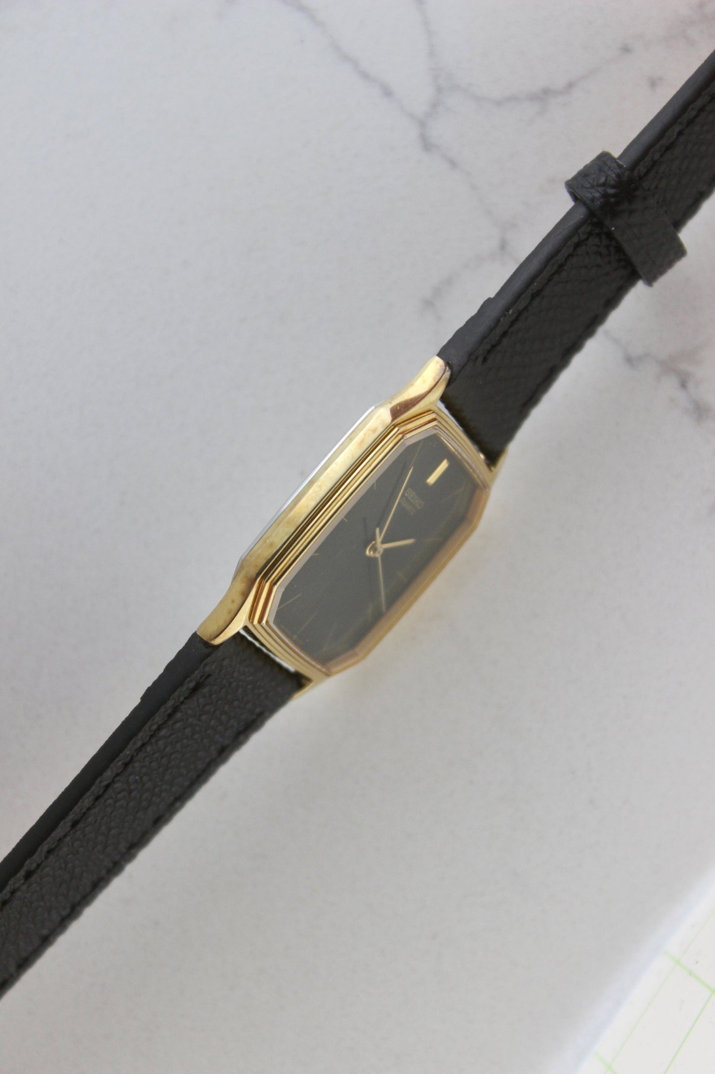 A close-up of the Seiko Chariot 9021-5180
featuring a sleek rectangular gold-plated case, a black dial with minimalist gold indices, and delicate gold-tone hands. The modern yet timeless design is paired with a handmade black leather strap stitched with beige accents for a refined look.