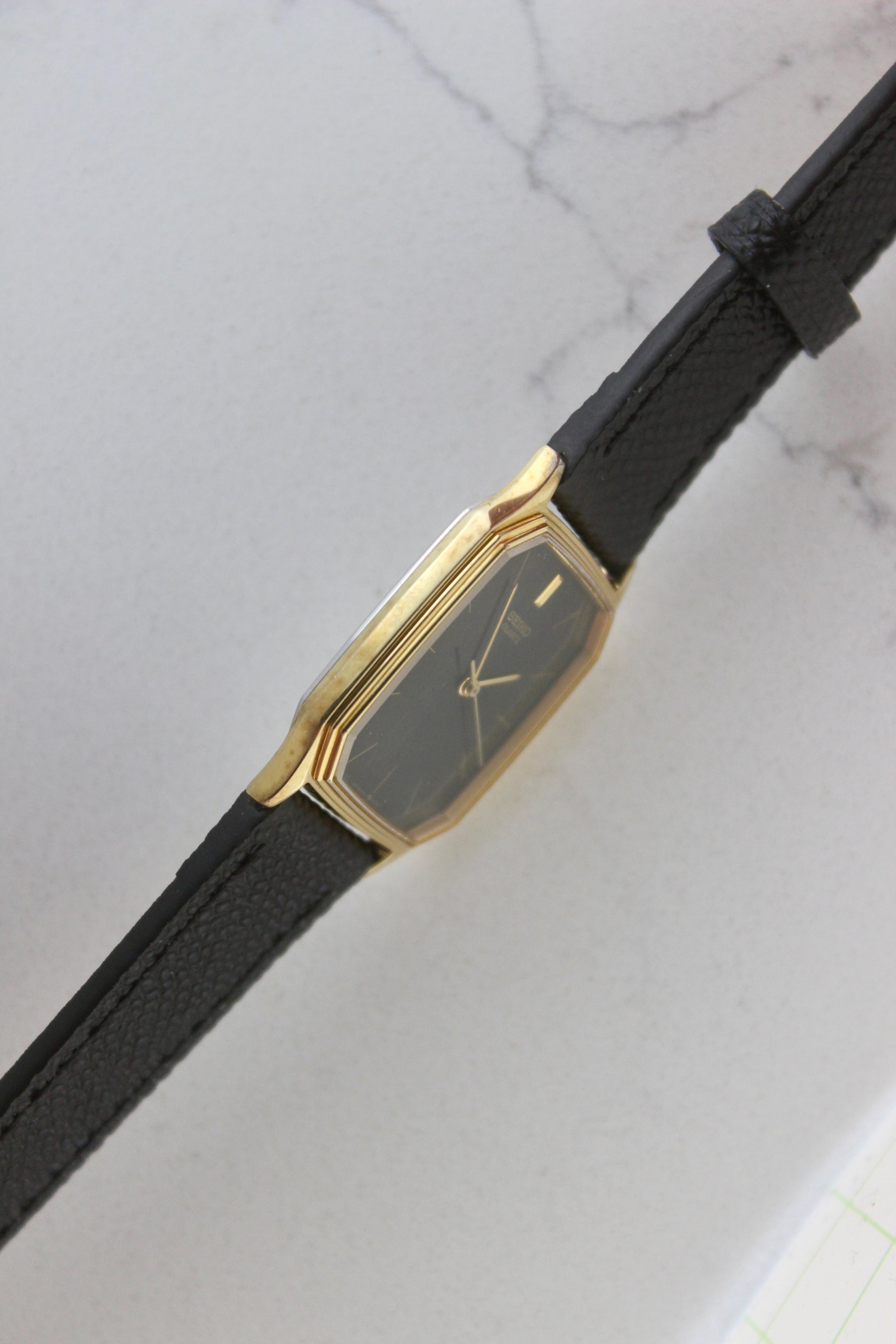 A close-up of the Seiko Chariot 9021-5180
featuring a sleek rectangular gold-plated case, a black dial with minimalist gold indices, and delicate gold-tone hands. The modern yet timeless design is paired with a handmade black leather strap stitched with beige accents for a refined look.