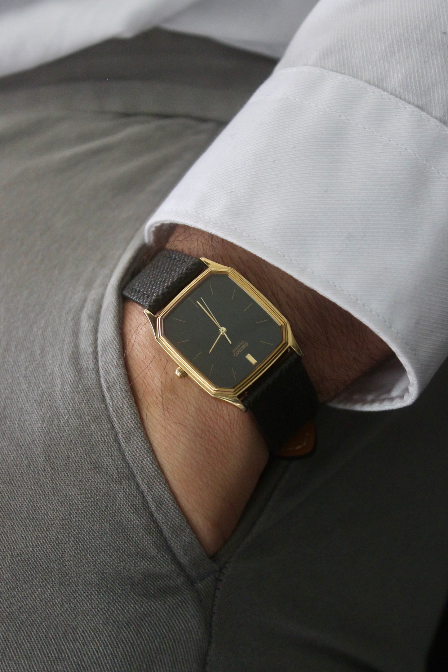 A close-up of the Seiko Chariot 69021-5180
featuring a sleek rectangular gold-plated case, a black dial with minimalist gold indices, and delicate gold-tone hands. The modern yet timeless design is paired with a handmade black leather strap stitched with beige accents for a refined look.