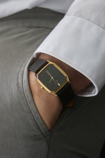 A close-up of the Seiko Chariot 69021-5180
featuring a sleek rectangular gold-plated case, a black dial with minimalist gold indices, and delicate gold-tone hands. The modern yet timeless design is paired with a handmade black leather strap stitched with beige accents for a refined look.