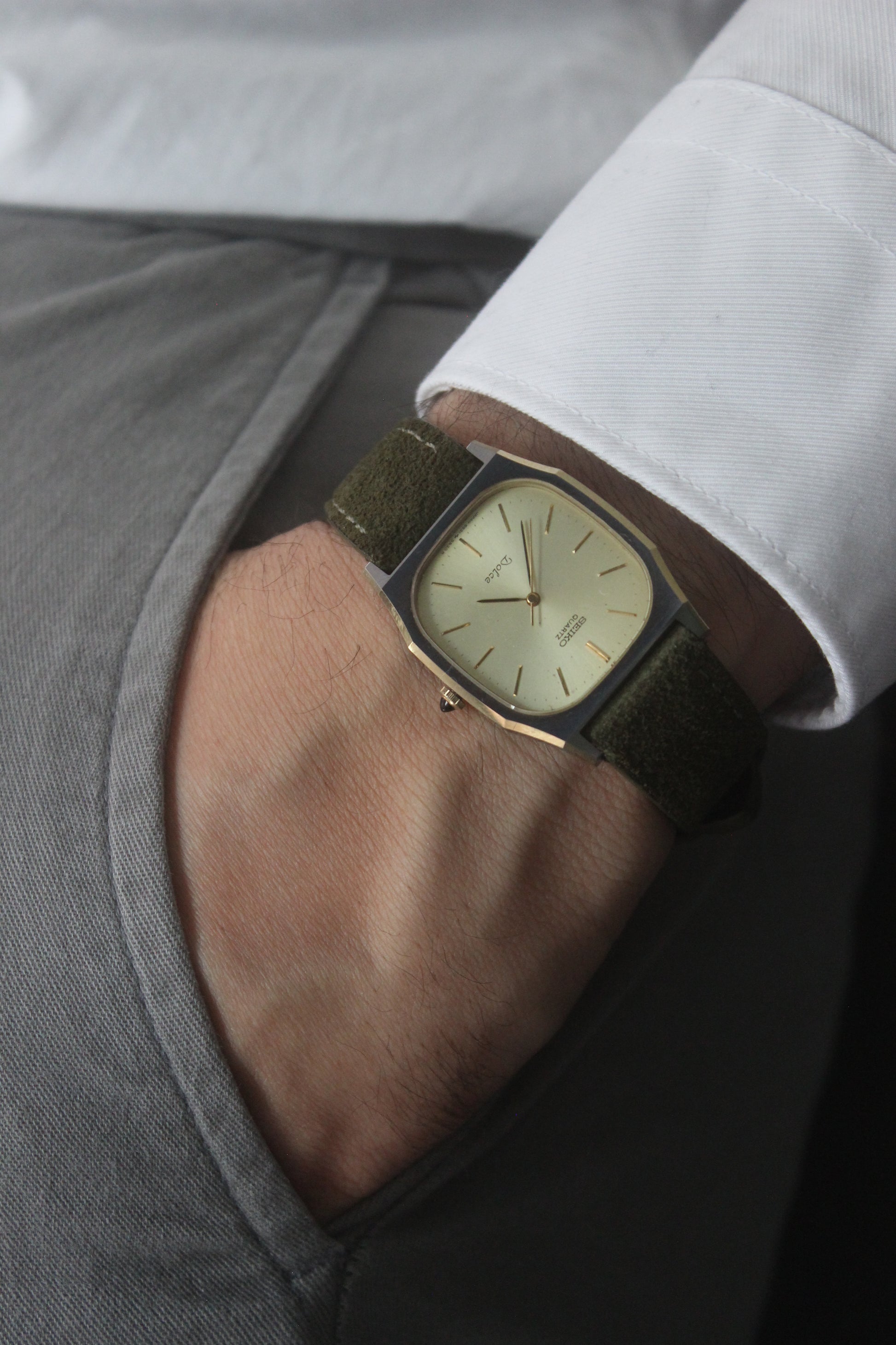 A close-up of the Seiko Dolce 5931-5420
watch featuring a rectangular stainless steel case with a gold-plated bezel, a champagne dial, and gold hour indices. Paired with an elegant green suede strap, this minimalist design showcases timeless sophistication.
