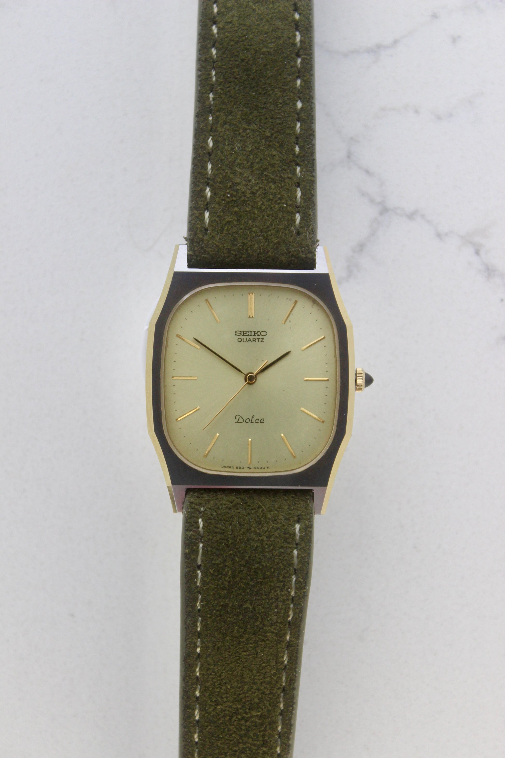 A close-up of the Seiko Dolce 5931-5420
watch featuring a rectangular stainless steel case with a gold-plated bezel, a champagne dial, and gold hour indices. Paired with an elegant green suede strap, this minimalist design showcases timeless sophistication.