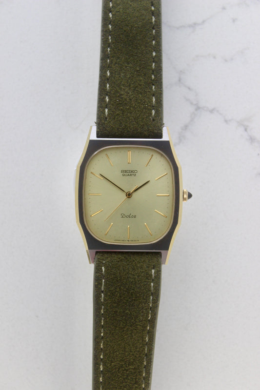 A close-up of the Seiko Dolce 5931-5420
watch featuring a rectangular stainless steel case with a gold-plated bezel, a champagne dial, and gold hour indices. Paired with an elegant green suede strap, this minimalist design showcases timeless sophistication.