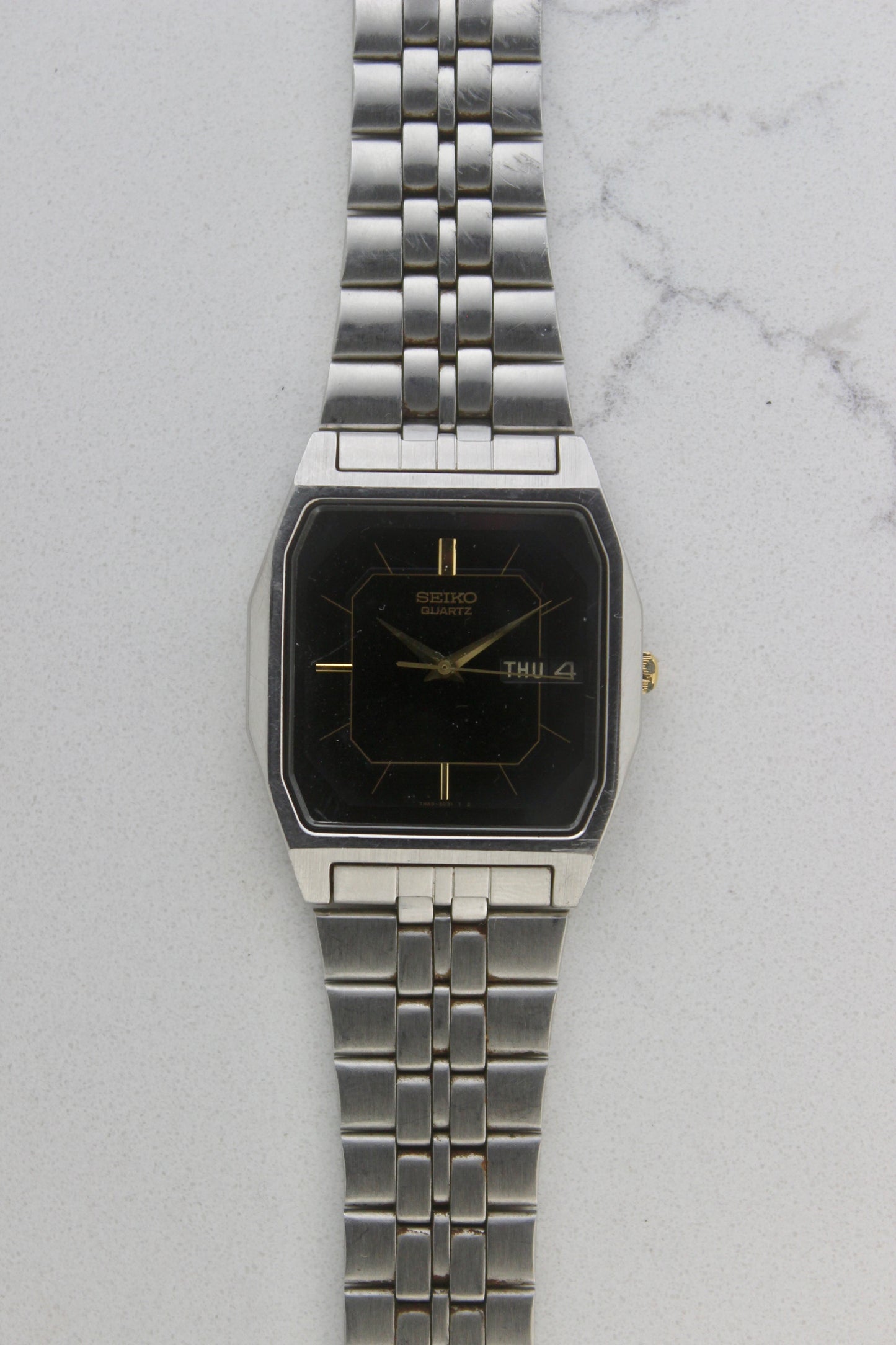 A close-up of the Seiko Dolce Quartz 7N43-5011, featuring a rectangular stainless steel case, black dial with gold accents, and a day-date window at 3 o'clock. The watch is paired with a stainless steel bracelet, exuding a timeless and versatile design.