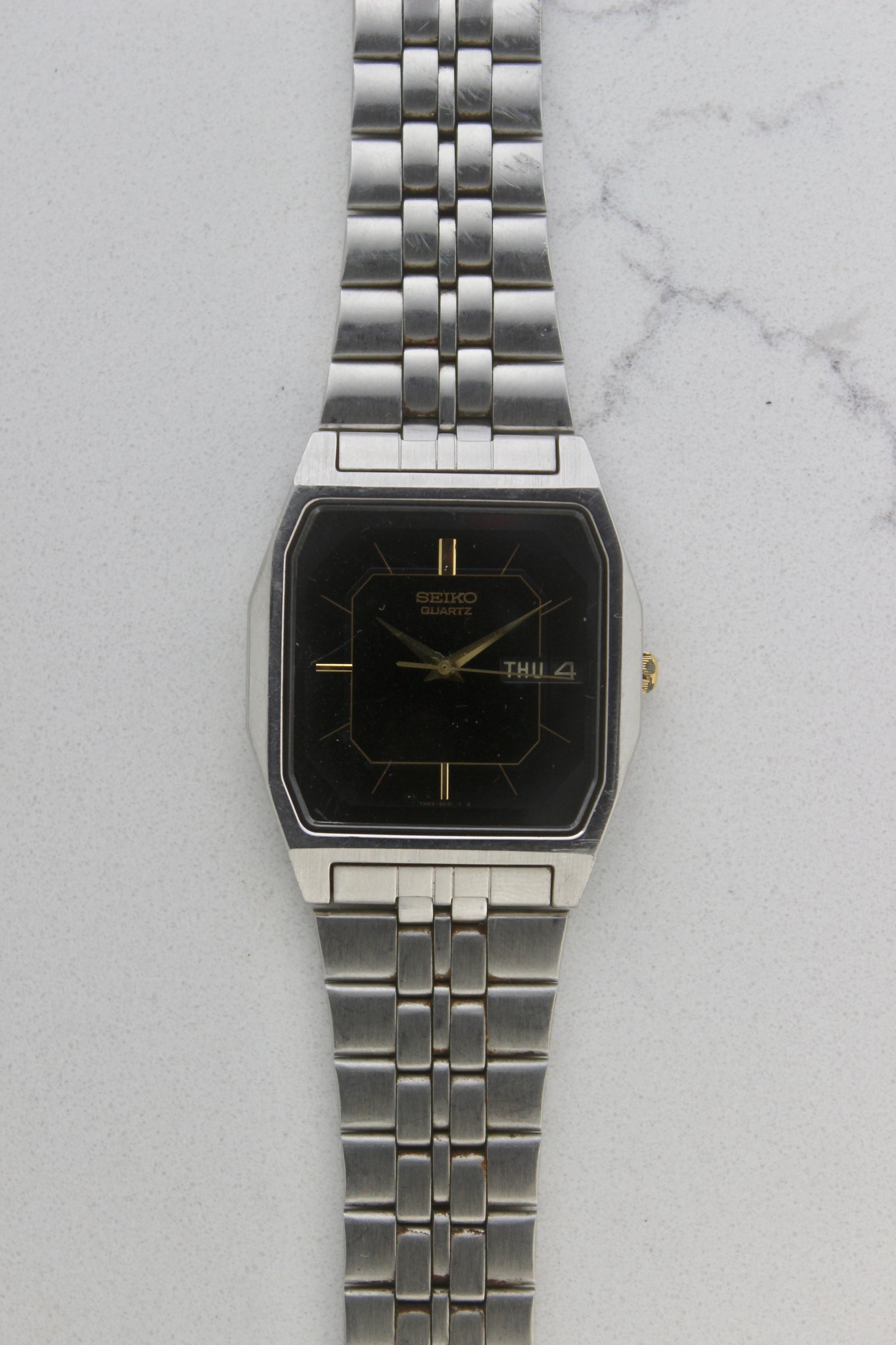 A close-up of the Seiko Dolce Quartz 7N43-5011, featuring a rectangular stainless steel case, black dial with gold accents, and a day-date window at 3 o'clock. The watch is paired with a stainless steel bracelet, exuding a timeless and versatile design.