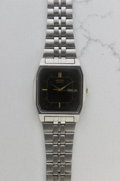 A close-up of the Seiko Dolce Quartz 7N43-5011, featuring a rectangular stainless steel case, black dial with gold accents, and a day-date window at 3 o'clock. The watch is paired with a stainless steel bracelet, exuding a timeless and versatile design.