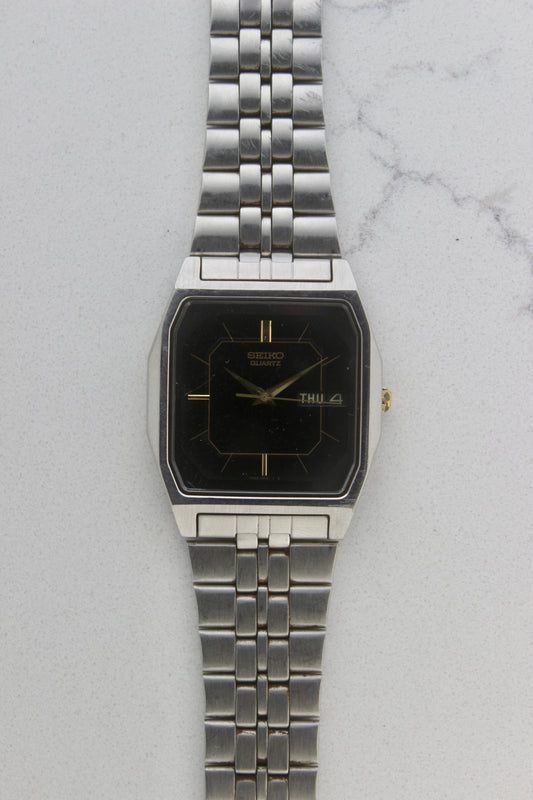 A close-up of the Seiko Dolce Quartz 7N43-5011, featuring a rectangular stainless steel case, black dial with gold accents, and a day-date window at 3 o'clock. The watch is paired with a stainless steel bracelet, exuding a timeless and versatile design.