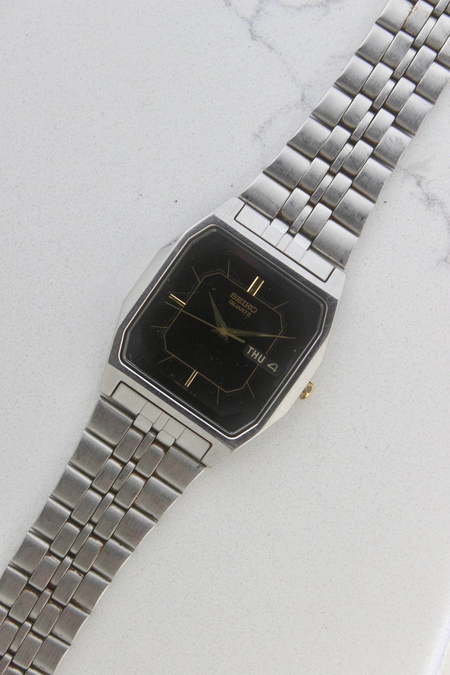 A close-up of the Seiko Dolce Quartz 7N43-5011, featuring a rectangular stainless steel case, black dial with gold accents, and a day-date window at 3 o'clock. The watch is paired with a stainless steel bracelet, exuding a timeless and versatile design.