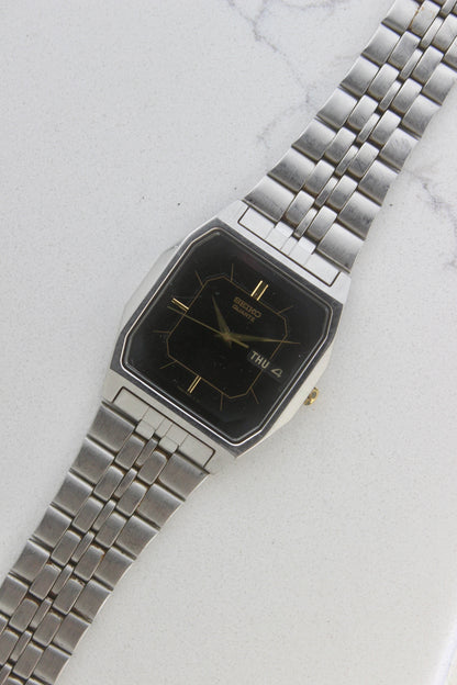 A close-up of the Seiko Dolce Quartz 7N43-5011, featuring a rectangular stainless steel case, black dial with gold accents, and a day-date window at 3 o'clock. The watch is paired with a stainless steel bracelet, exuding a timeless and versatile design.