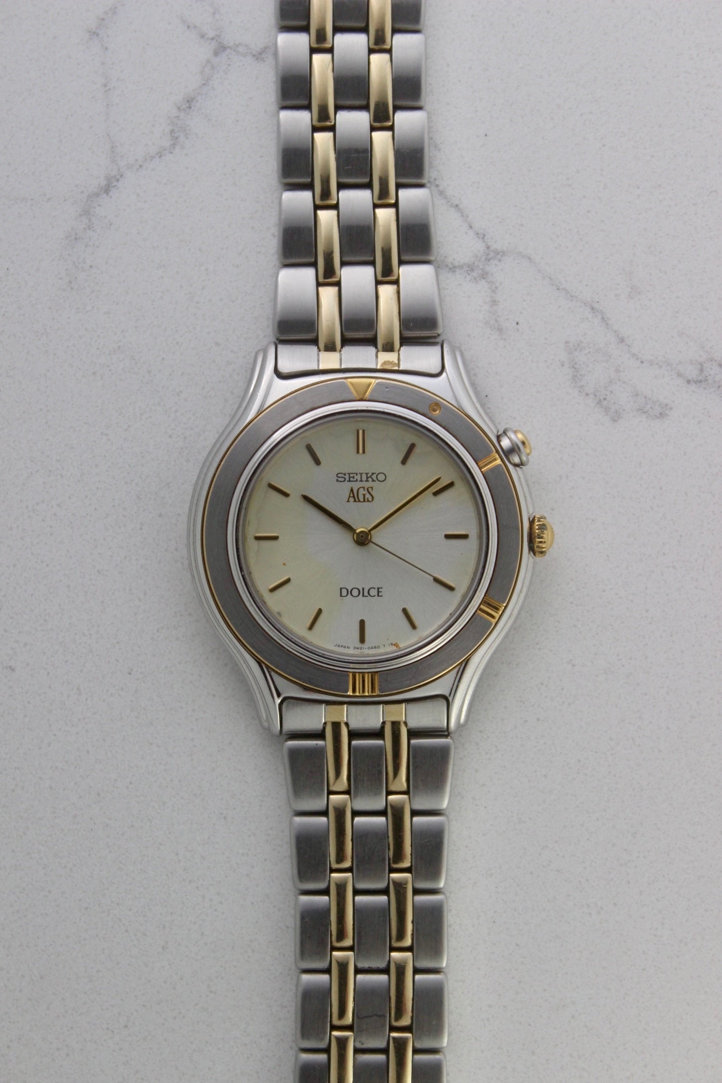 Seiko Dolce AGS wristwatch with a two-tone stainless steel and gold-plated bracelet, set against a light-colored marble background. The watch features a pearl white sunburst dial with gold-plated stick indices and hands. The fixed bezel is stainless steel with gold accents. The case measures approximately 37mm in diameter and 41mm lug to lug. The dial includes the text
'Seiko AGS' at the top and 'Dolce' at the bottom. A gold-toned crown is positioned at 3 o'clock. 