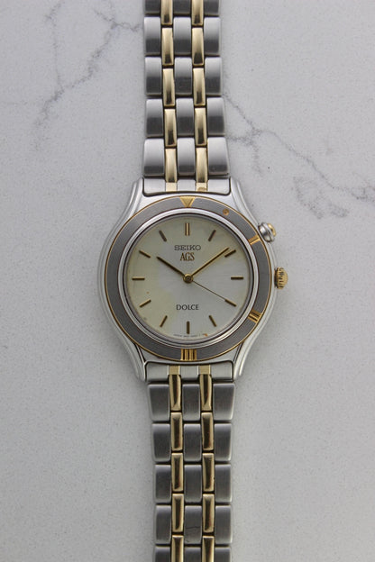 Seiko Dolce AGS wristwatch with a two-tone stainless steel and gold-plated bracelet, set against a light-colored marble background. The watch features a pearl white sunburst dial with gold-plated stick indices and hands. The fixed bezel is stainless steel with gold accents. The case measures approximately 37mm in diameter and 41mm lug to lug. The dial includes the text
'Seiko AGS' at the top and 'Dolce' at the bottom. A gold-toned crown is positioned at 3 o'clock. 