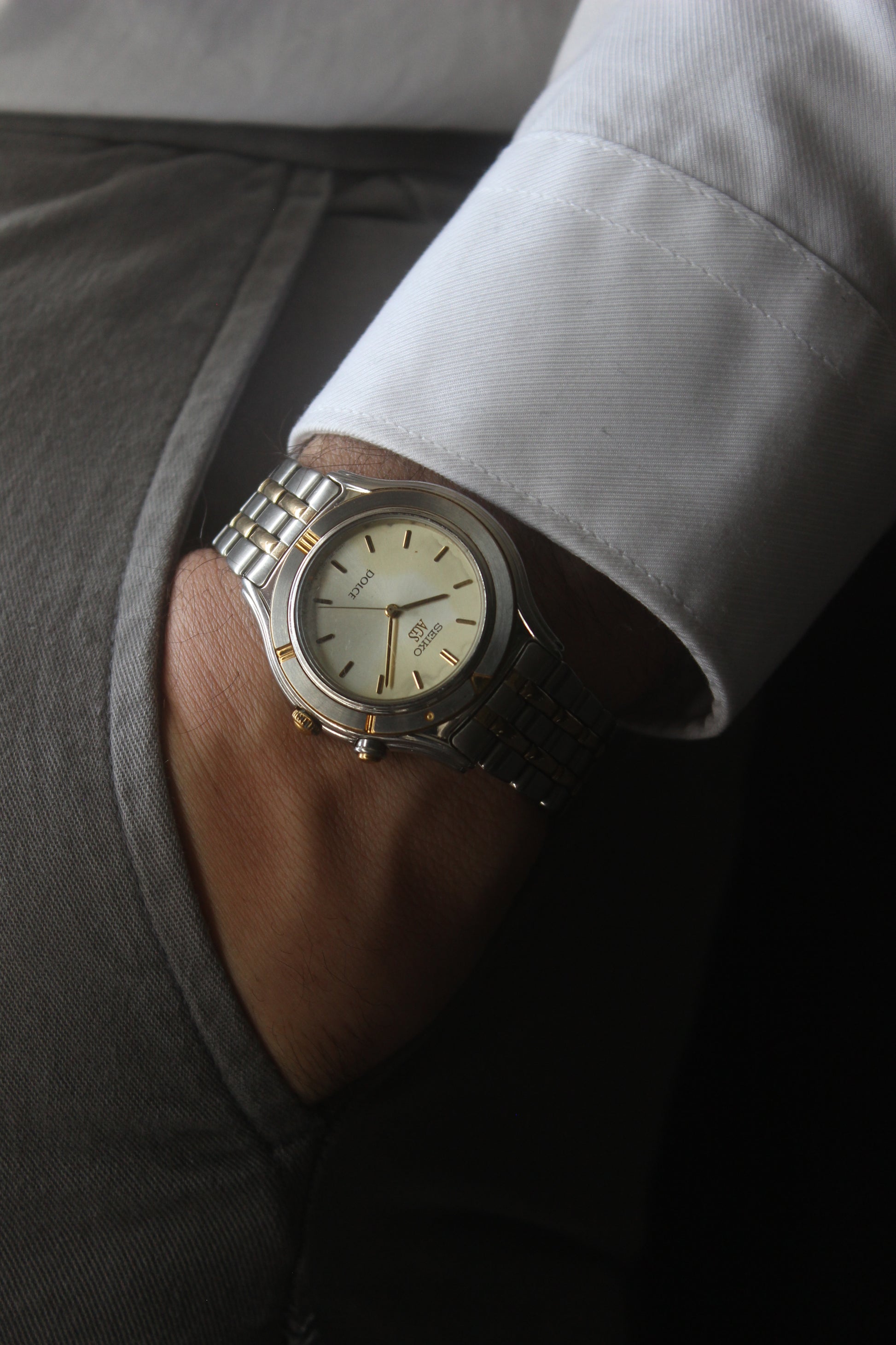 vintage Seiko Dolce AGS wristwatch with a two-tone stainless steel and gold-plated bracelet, set against a light-colored marble background. The watch features a pearl white sunburst dial with gold-plated stick indices and hands. The fixed bezel is stainless steel with gold accents. The case measures approximately 37mm in diameter and 41mm lug to lug. The dial includes the text
'Seiko AGS' at the top and 'Dolce' at the bottom. A gold-toned crown is positioned at 3 o'clock. 