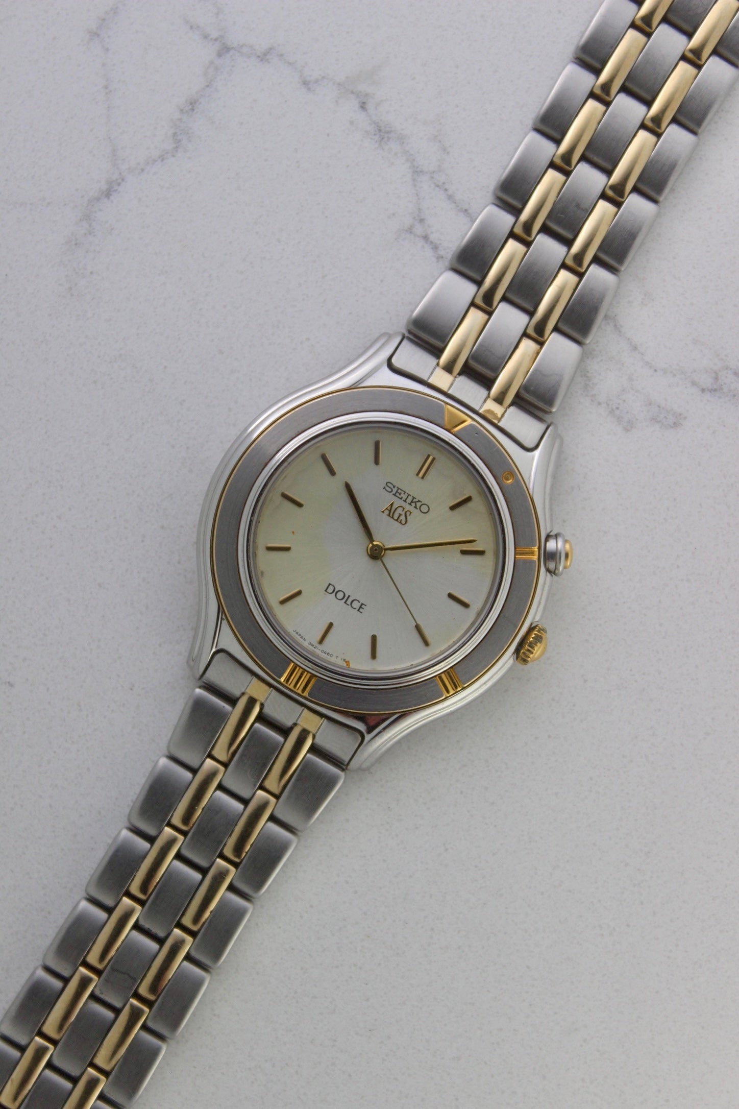 vintage Seiko Dolce AGS wristwatch with a two-tone stainless steel and gold-plated bracelet, set against a light-colored marble background. The watch features a pearl white sunburst dial with gold-plated stick indices and hands. The fixed bezel is stainless steel with gold accents. The case measures approximately 37mm in diameter and 41mm lug to lug. The dial includes the text
'Seiko AGS' at the top and 'Dolce' at the bottom. A gold-toned crown is positioned at 3 o'clock. 
