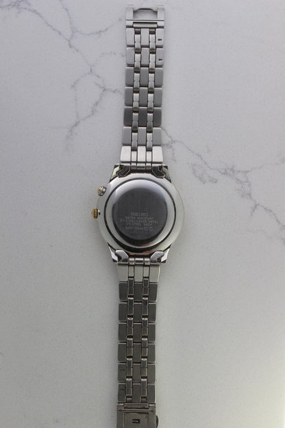 vintage Seiko Dolce AGS wristwatch with a two-tone stainless steel and gold-plated bracelet, set against a light-colored marble background. The watch features a pearl white sunburst dial with gold-plated stick indices and hands. The fixed bezel is stainless steel with gold accents. The case measures approximately 37mm in diameter and 41mm lug to lug. The dial includes the text
'Seiko AGS' at the top and 'Dolce' at the bottom. A gold-toned crown is positioned at 3 o'clock. 