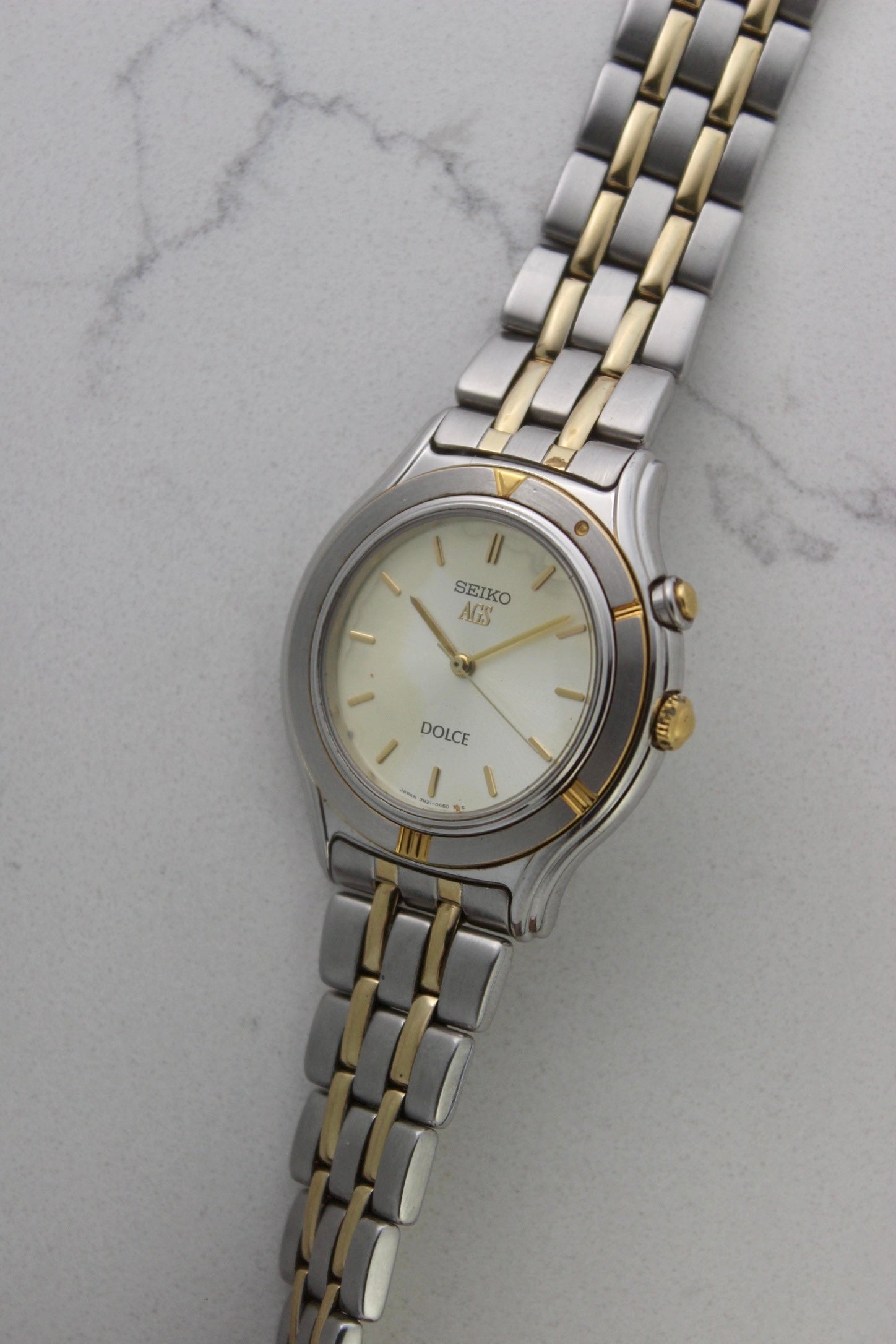 vintage Seiko Dolce AGS wristwatch with a two-tone stainless steel and gold-plated bracelet, set against a light-colored marble background. The watch features a pearl white sunburst dial with gold-plated stick indices and hands. The fixed bezel is stainless steel with gold accents. The case measures approximately 37mm in diameter and 41mm lug to lug. The dial includes the text
'Seiko AGS' at the top and 'Dolce' at the bottom. A gold-toned crown is positioned at 3 o'clock. 
