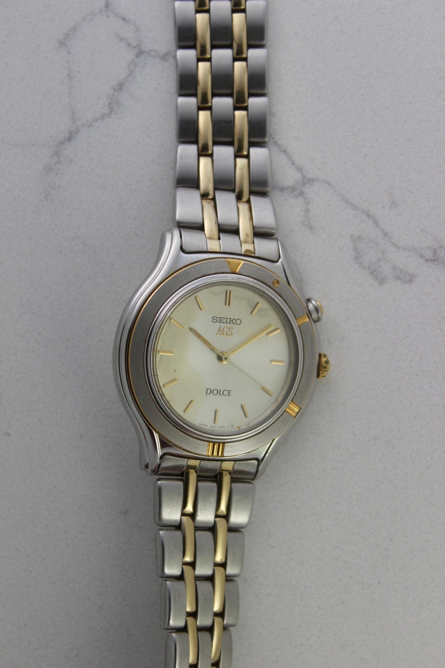 vintage Seiko Dolce AGS wristwatch with a two-tone stainless steel and gold-plated bracelet, set against a light-colored marble background. The watch features a pearl white sunburst dial with gold-plated stick indices and hands. The fixed bezel is stainless steel with gold accents. The case measures approximately 37mm in diameter and 41mm lug to lug. The dial includes the text
'Seiko AGS' at the top and 'Dolce' at the bottom. A gold-toned crown is positioned at 3 o'clock. 