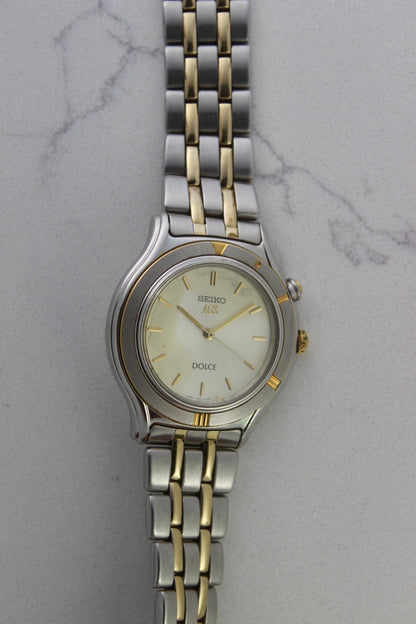 vintage Seiko Dolce AGS wristwatch with a two-tone stainless steel and gold-plated bracelet, set against a light-colored marble background. The watch features a pearl white sunburst dial with gold-plated stick indices and hands. The fixed bezel is stainless steel with gold accents. The case measures approximately 37mm in diameter and 41mm lug to lug. The dial includes the text
'Seiko AGS' at the top and 'Dolce' at the bottom. A gold-toned crown is positioned at 3 o'clock. 
