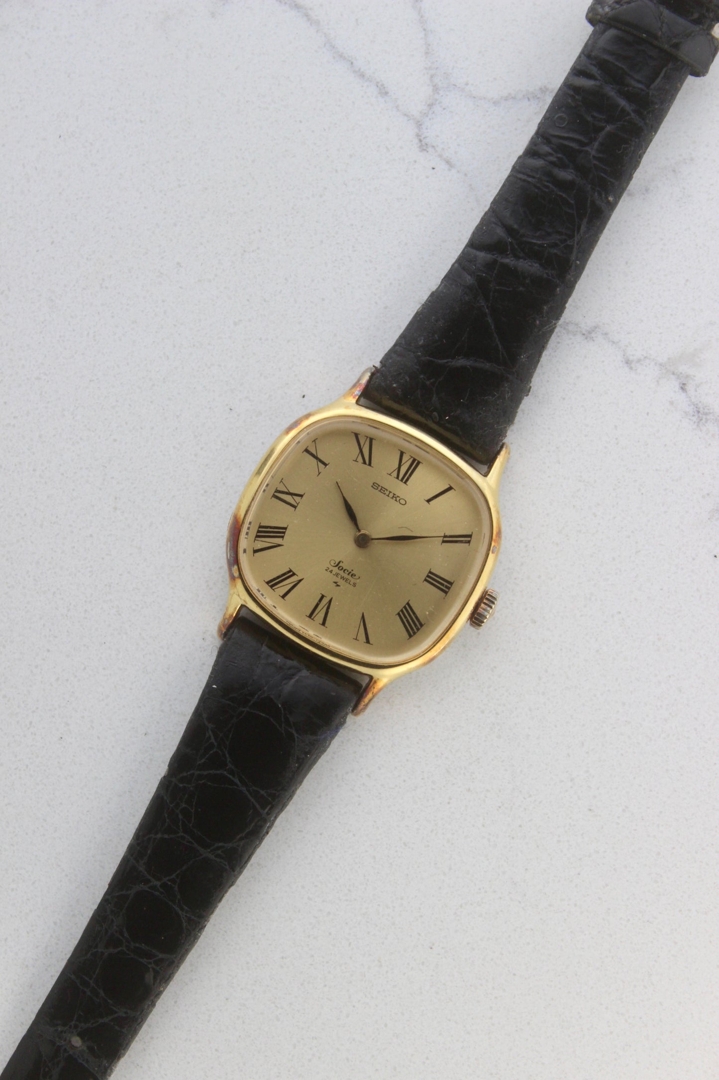 A close-up of the Seiko Socie Ref.
2220-3470, featuring a cushion-shaped gold-filled case, a champagne dial with black Roman numeral markers, and sleek black hands. Paired with a black leather strap, this vintage timepiece exudes elegance and timeless style.