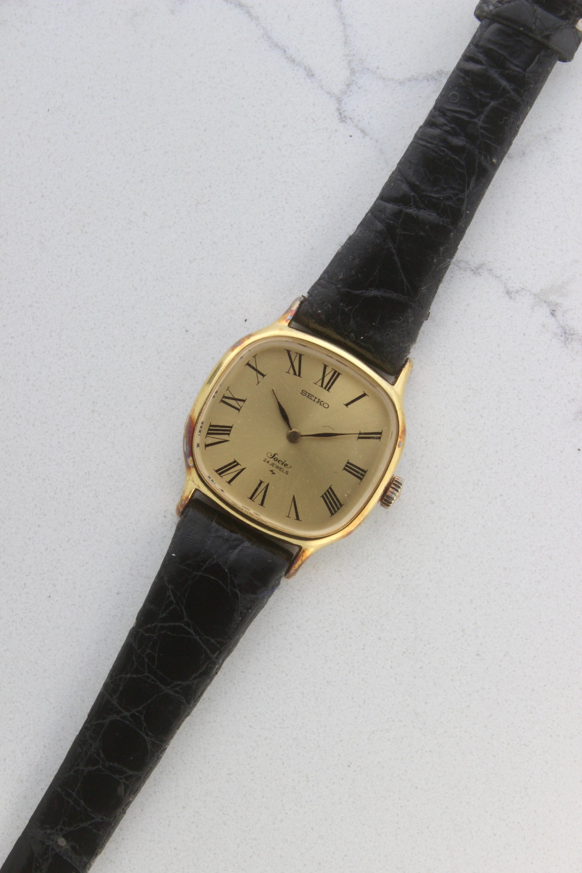 A close-up of the Seiko Socie Ref.
2220-3470, featuring a cushion-shaped gold-filled case, a champagne dial with black Roman numeral markers, and sleek black hands. Paired with a black leather strap, this vintage timepiece exudes elegance and timeless style.