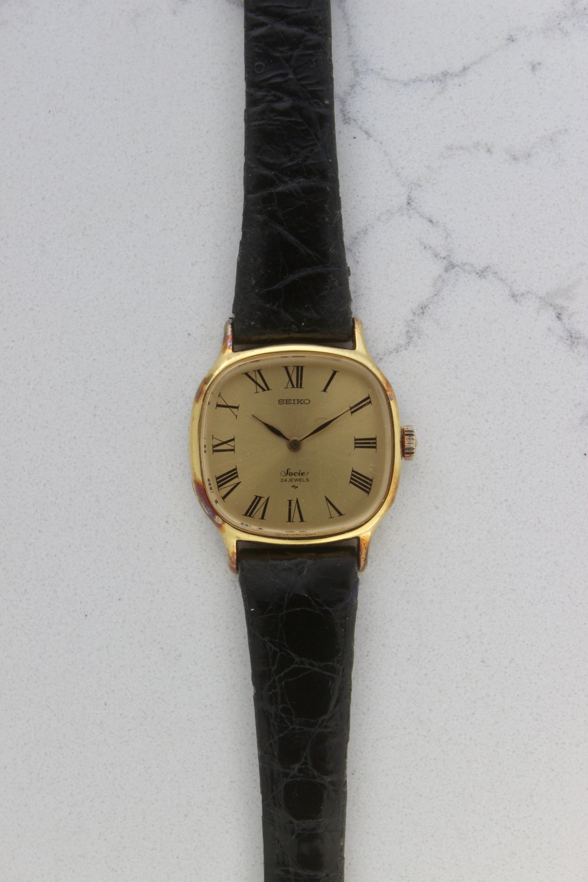 A close-up of the Seiko Socie Ref.
2220-3470, featuring a cushion-shaped gold-filled case, a champagne dial with black Roman numeral markers, and sleek black hands. Paired with a black leather strap, this vintage timepiece exudes elegance and timeless style.