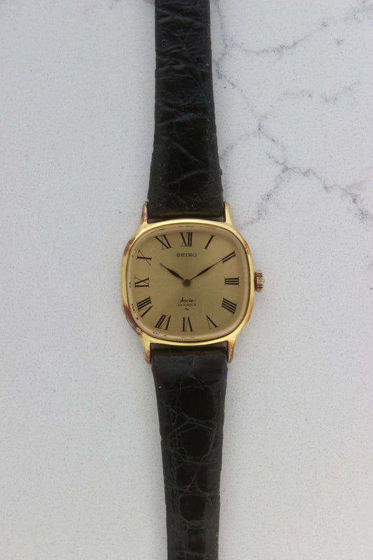 A close-up of the Seiko Socie Ref.
2220-3470, featuring a cushion-shaped gold-filled case, a champagne dial with black Roman numeral markers, and sleek black hands. Paired with a black leather strap, this vintage timepiece exudes elegance and timeless style.