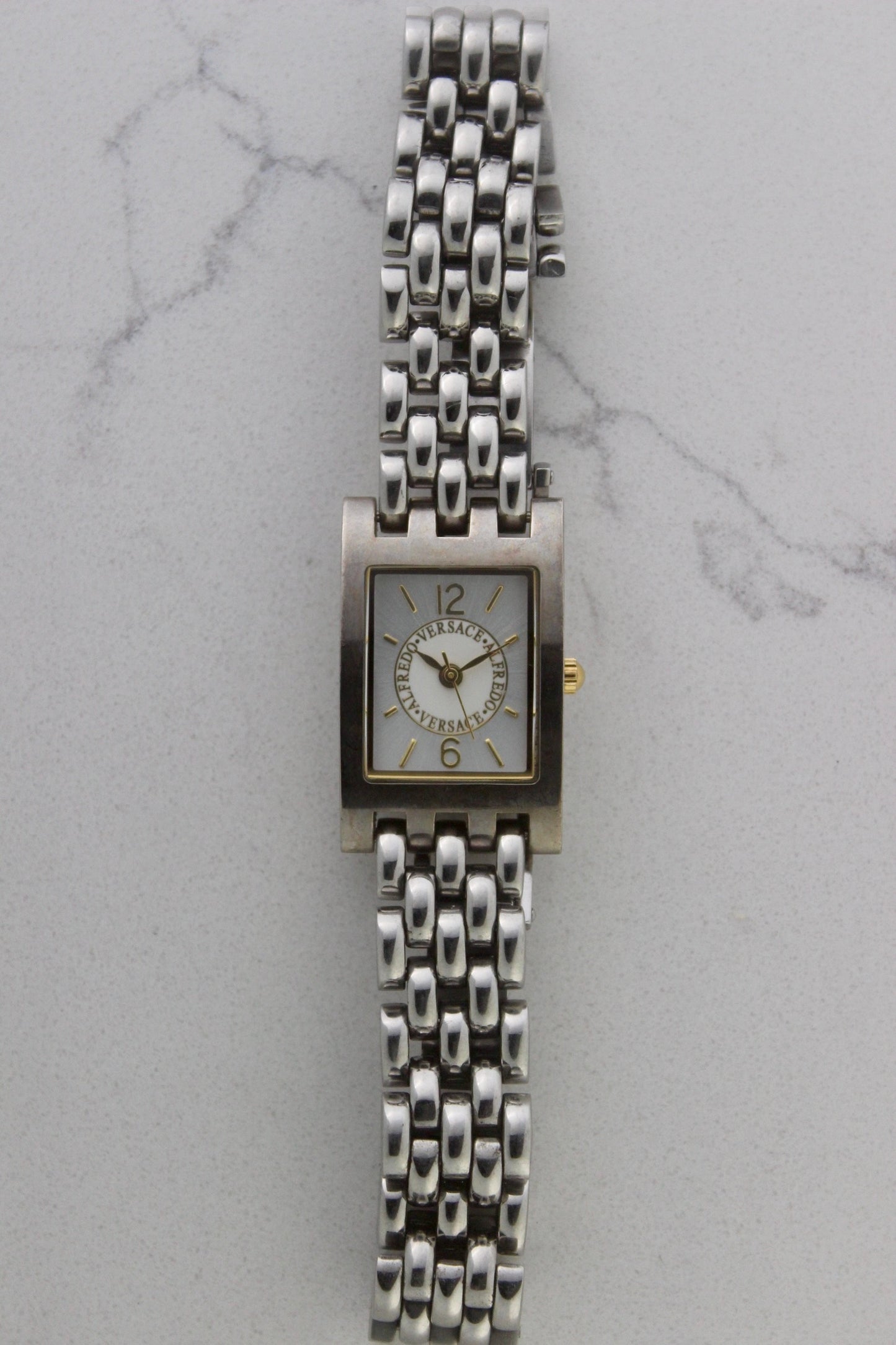 a rectangular stainless steel wristwatch with a silver-tone case and bracelet featuring a multi-link design. The watch face has a while dial with gold-tone hour makers, hands, and a cirular logo pattern that reads 'ALFREDO VERSACE'. The crown is gold-tone and positioned on the right side of the case. The watch is displayed against a light marble surface background. 