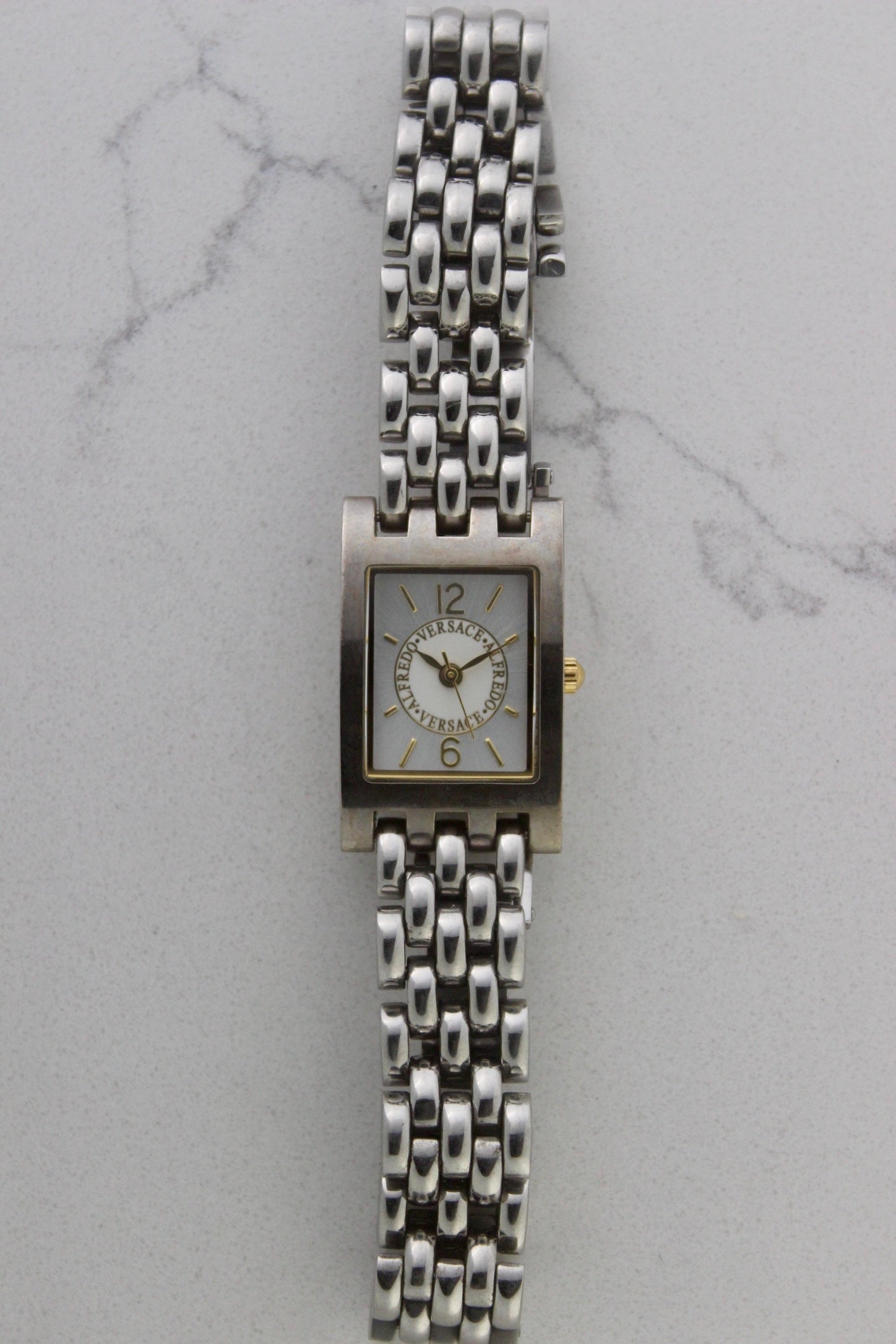 a rectangular stainless steel wristwatch with a silver-tone case and bracelet featuring a multi-link design. The watch face has a while dial with gold-tone hour makers, hands, and a cirular logo pattern that reads 'ALFREDO VERSACE'. The crown is gold-tone and positioned on the right side of the case. The watch is displayed against a light marble surface background. 