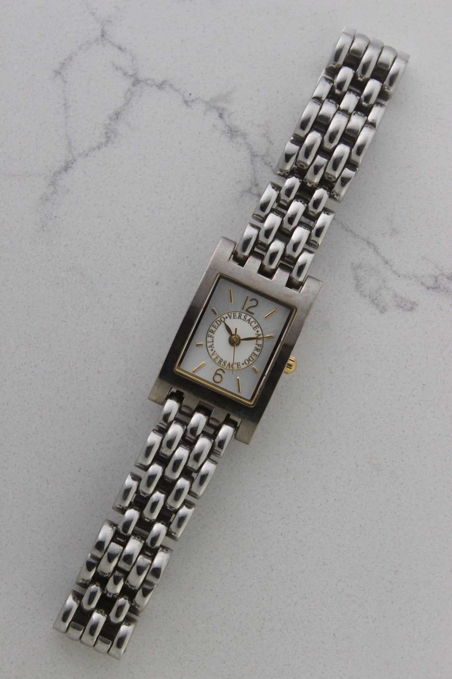 a rectangular stainless steel wristwatch with a silver-tone case and bracelet featuring a multi-link design. The watch face has a while dial with gold-tone hour makers, hands, and a cirular logo pattern that reads 'ALFREDO VERSACE'. The crown is gold-tone and positioned on the right side of the case. The watch is displayed against a light marble surface background. 