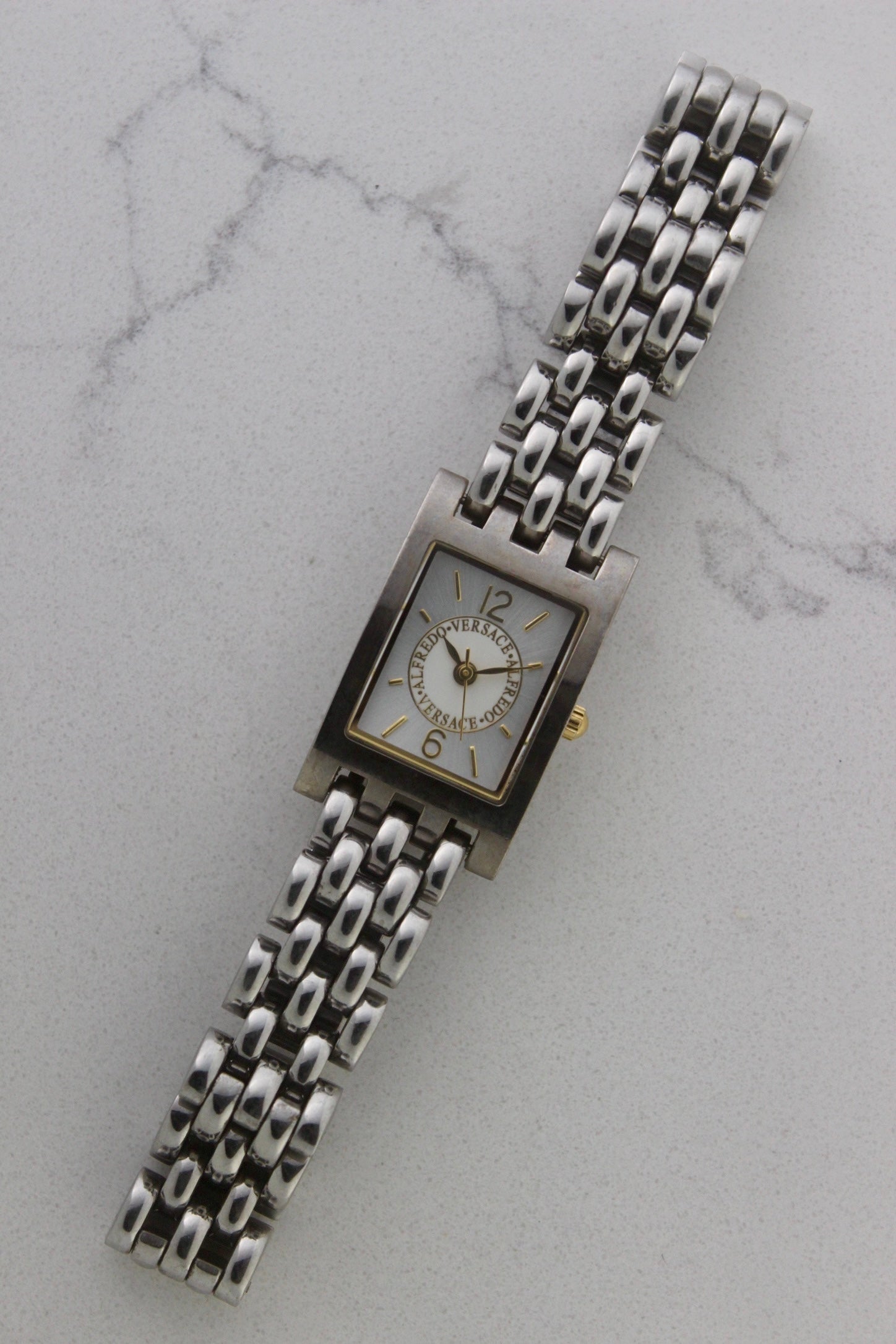 a rectangular stainless steel wristwatch with a silver-tone case and bracelet featuring a multi-link design. The watch face has a while dial with gold-tone hour makers, hands, and a cirular logo pattern that reads 'ALFREDO VERSACE'. The crown is gold-tone and positioned on the right side of the case. The watch is displayed against a light marble surface background. 
