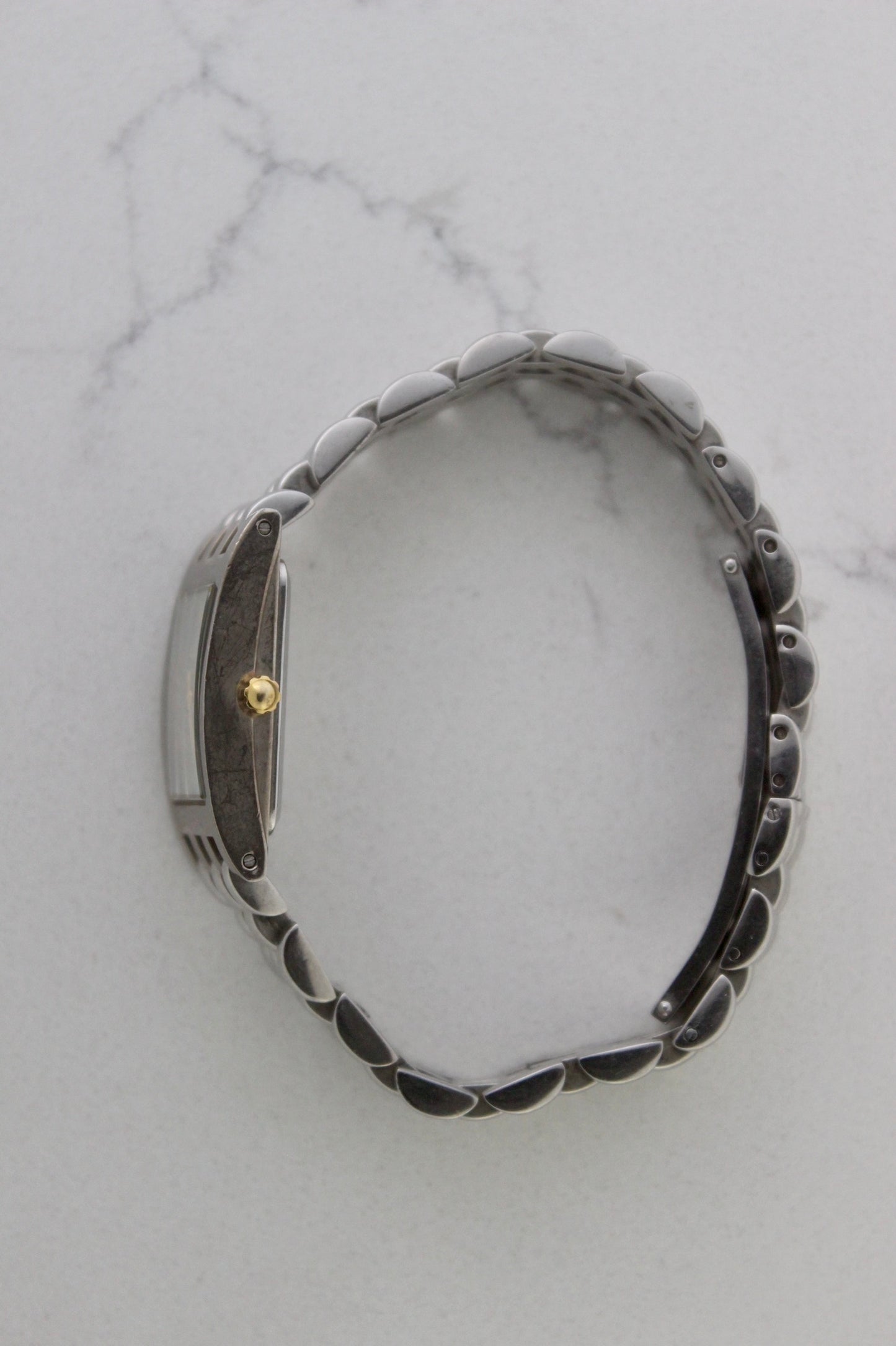 a rectangular stainless steel wristwatch with a silver-tone case and bracelet featuring a multi-link design. The watch face has a while dial with gold-tone hour makers, hands, and a cirular logo pattern that reads 'ALFREDO VERSACE'. The crown is gold-tone and positioned on the right side of the case. The watch is displayed against a light marble surface background. 