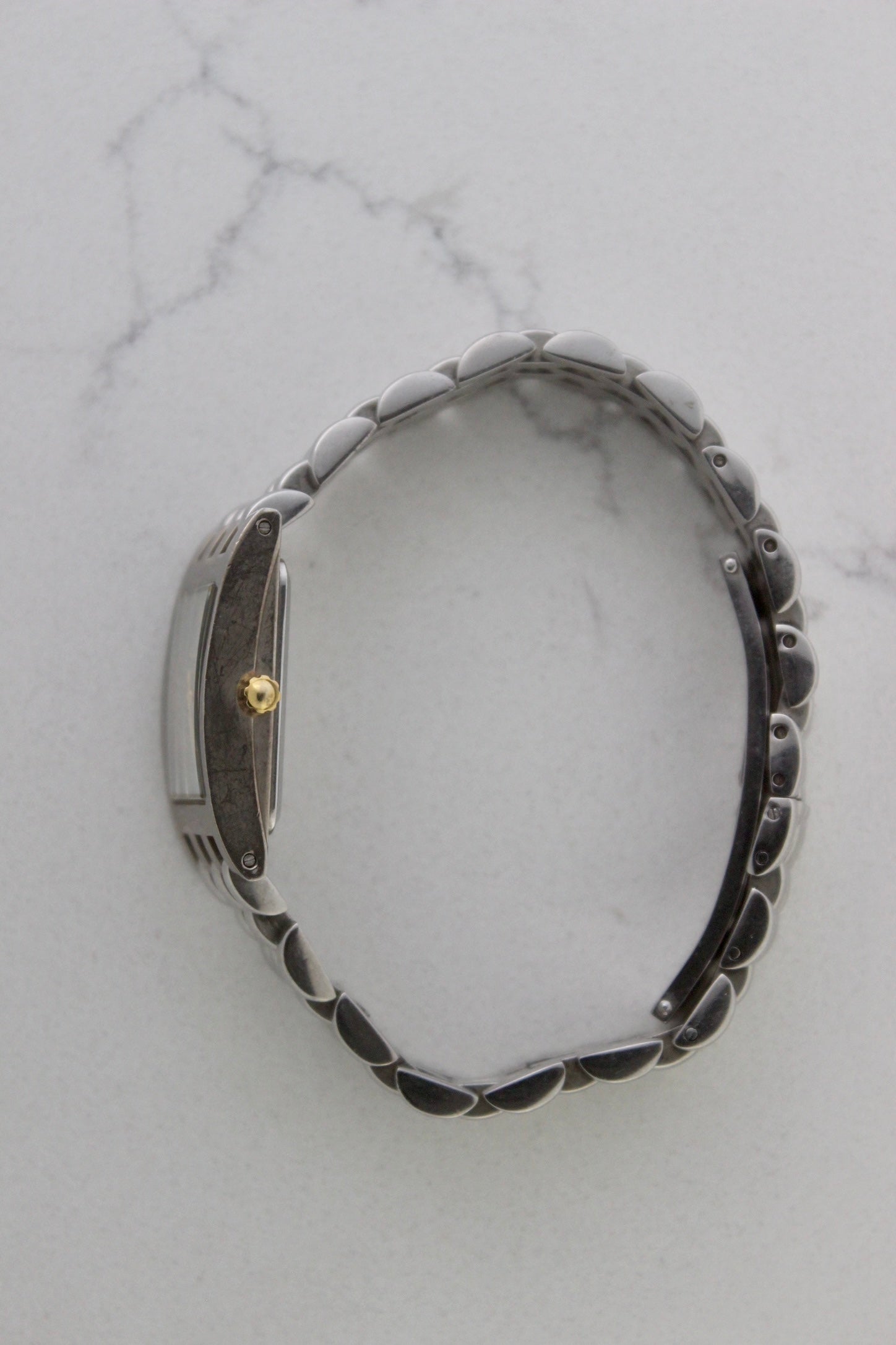 a rectangular stainless steel wristwatch with a silver-tone case and bracelet featuring a multi-link design. The watch face has a while dial with gold-tone hour makers, hands, and a cirular logo pattern that reads 'ALFREDO VERSACE'. The crown is gold-tone and positioned on the right side of the case. The watch is displayed against a light marble surface background. 