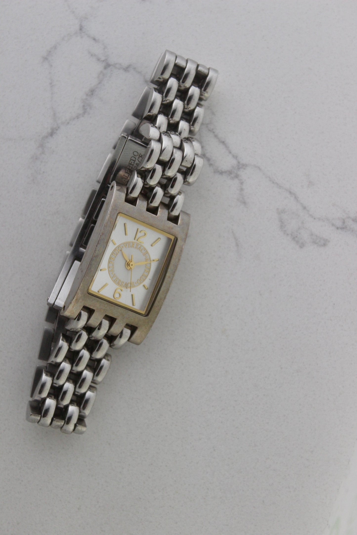 a rectangular stainless steel wristwatch with a silver-tone case and bracelet featuring a multi-link design. The watch face has a while dial with gold-tone hour makers, hands, and a cirular logo pattern that reads 'ALFREDO VERSACE'. The crown is gold-tone and positioned on the right side of the case. The watch is displayed against a light marble surface background. 