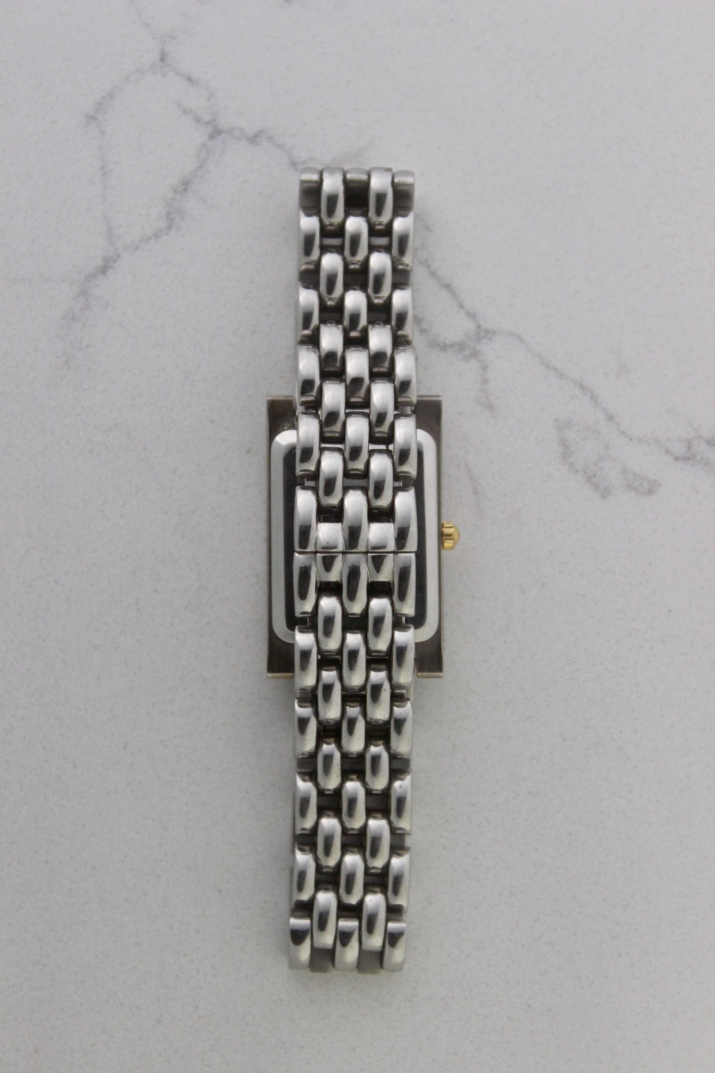 a rectangular stainless steel wristwatch with a silver-tone case and bracelet featuring a multi-link design. The watch face has a while dial with gold-tone hour makers, hands, and a cirular logo pattern that reads 'ALFREDO VERSACE'. The crown is gold-tone and positioned on the right side of the case. The watch is displayed against a light marble surface background. 