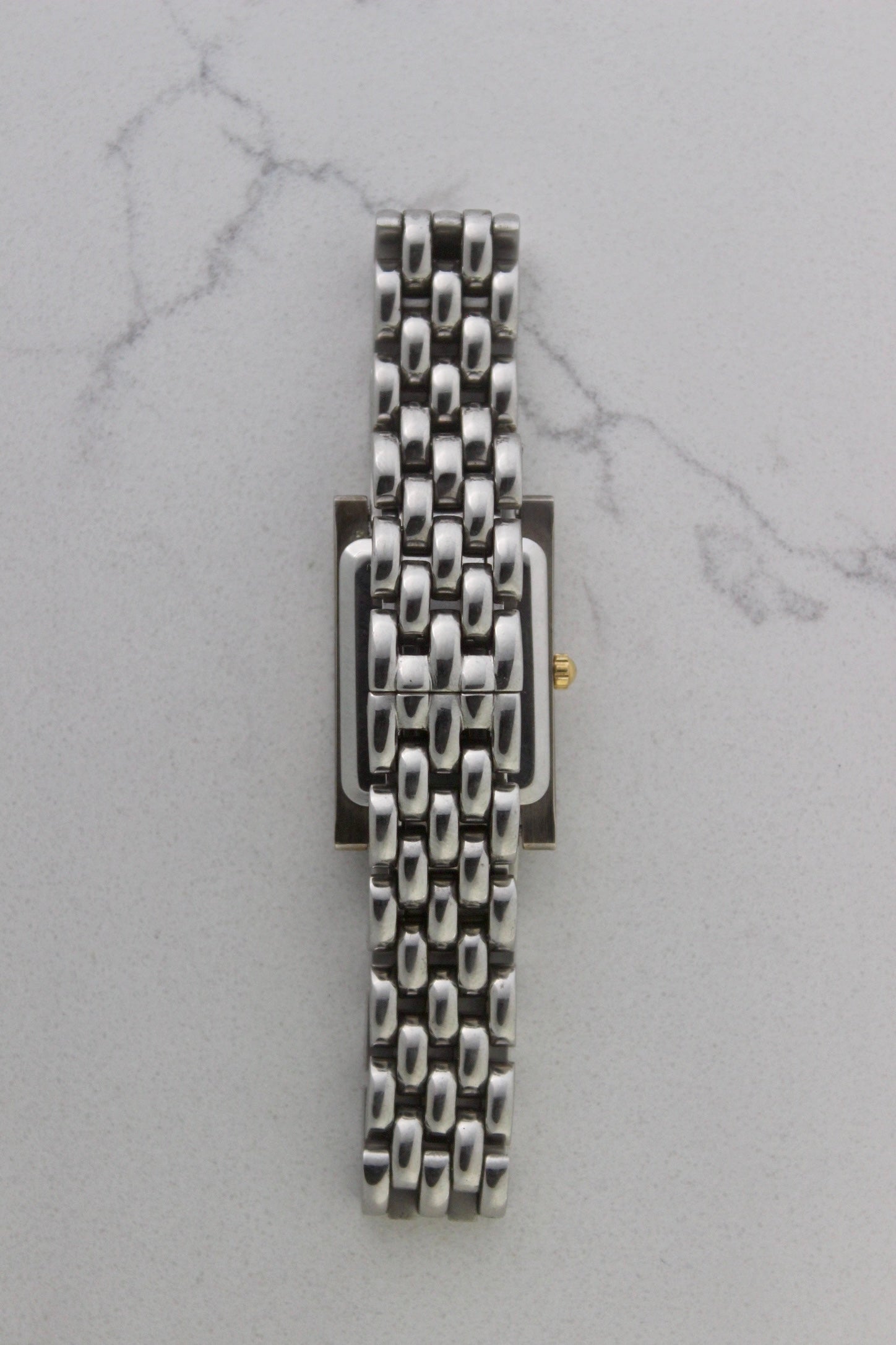 a rectangular stainless steel wristwatch with a silver-tone case and bracelet featuring a multi-link design. The watch face has a while dial with gold-tone hour makers, hands, and a cirular logo pattern that reads 'ALFREDO VERSACE'. The crown is gold-tone and positioned on the right side of the case. The watch is displayed against a light marble surface background. 