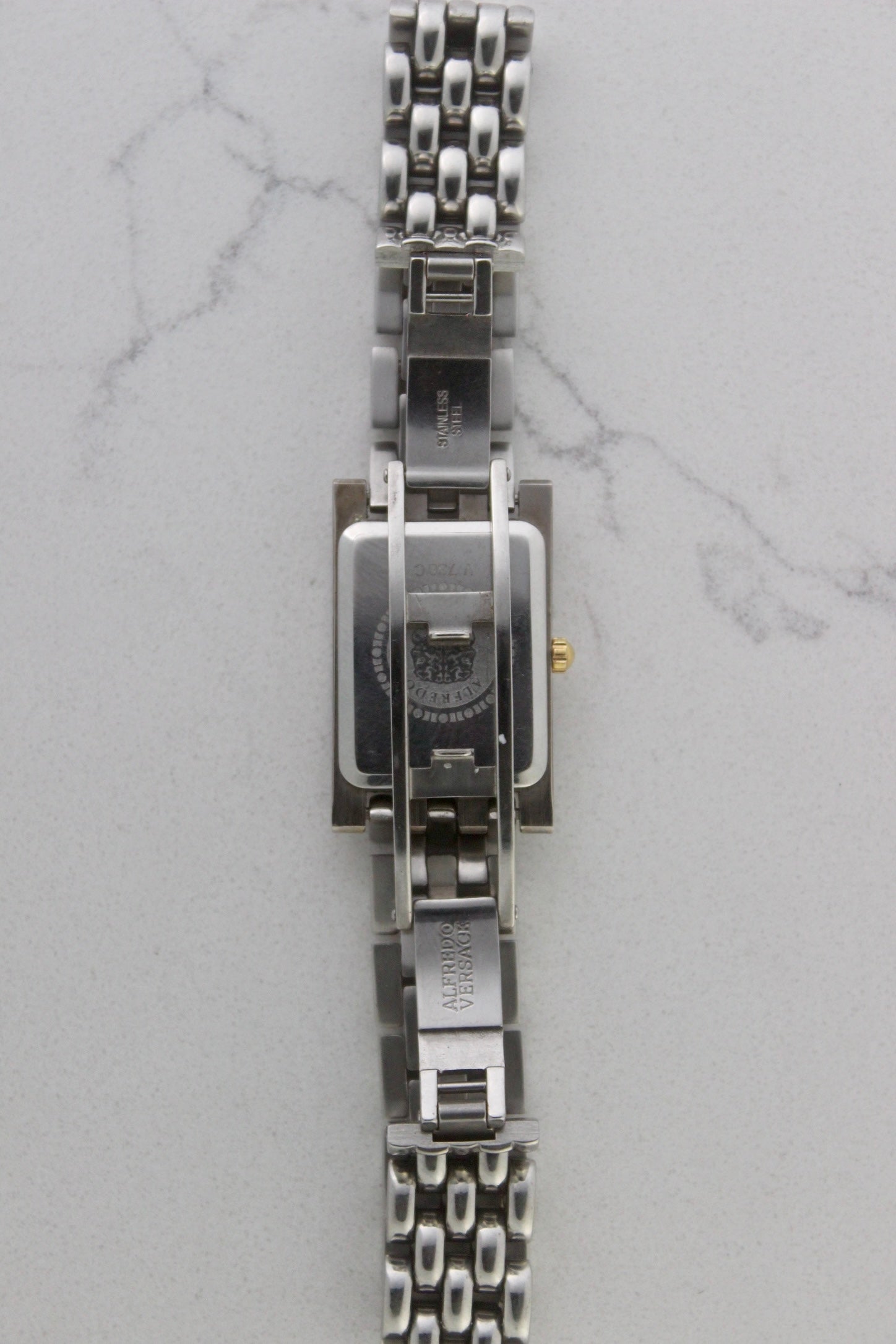 a rectangular stainless steel wristwatch with a silver-tone case and bracelet featuring a multi-link design. The watch face has a while dial with gold-tone hour makers, hands, and a cirular logo pattern that reads 'ALFREDO VERSACE'. The crown is gold-tone and positioned on the right side of the case. The watch is displayed against a light marble surface background. 