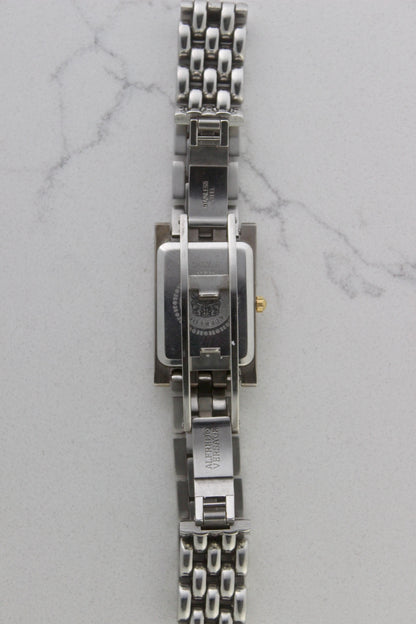 a rectangular stainless steel wristwatch with a silver-tone case and bracelet featuring a multi-link design. The watch face has a while dial with gold-tone hour makers, hands, and a cirular logo pattern that reads 'ALFREDO VERSACE'. The crown is gold-tone and positioned on the right side of the case. The watch is displayed against a light marble surface background. 